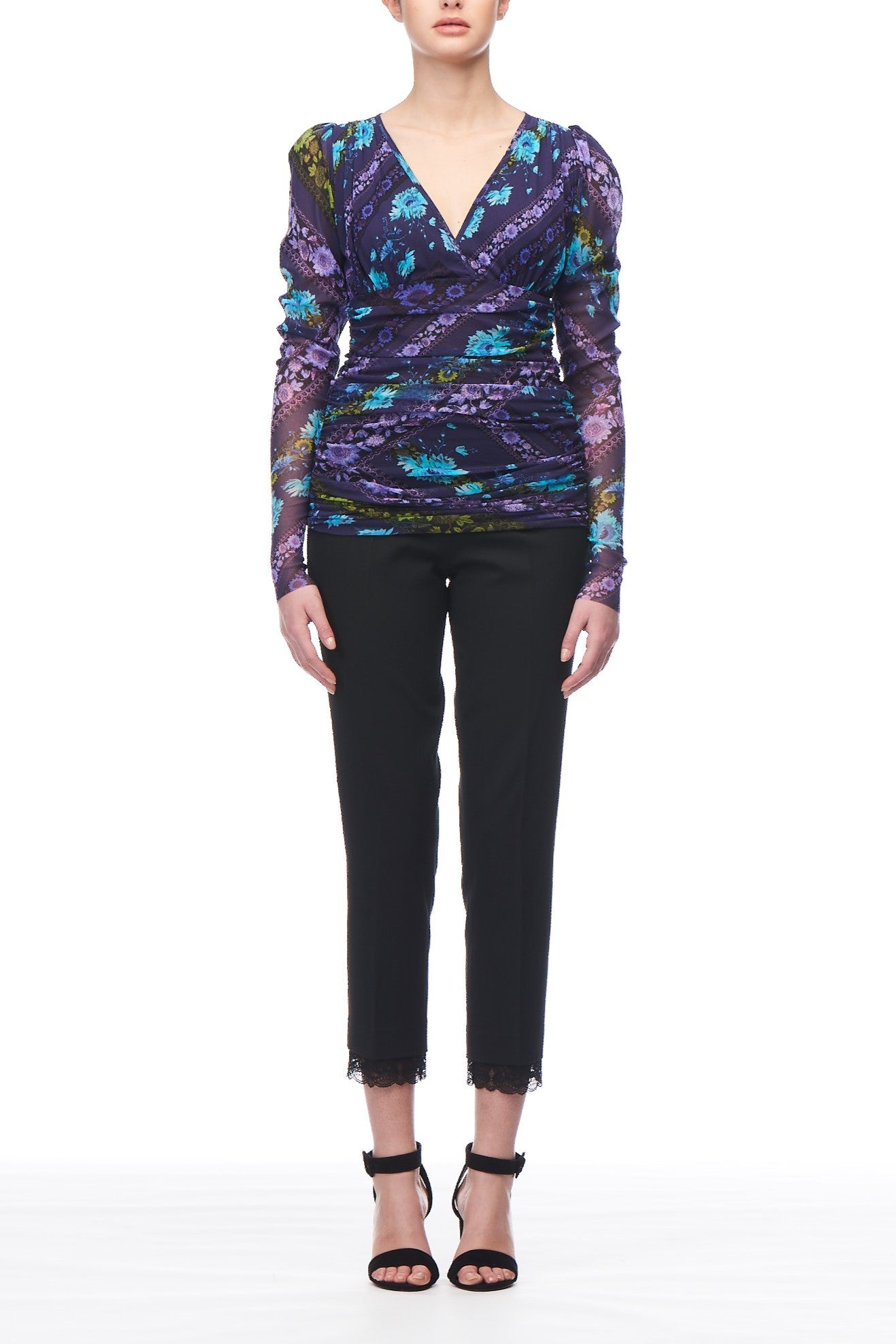 A person wearing the Fuzzi Woman Floral Mesh Top, adorned with romantic purple and blue floral patterns and ruched detailing, paired with black cropped pants featuring lace trim and black strappy heels, stands against a white background.