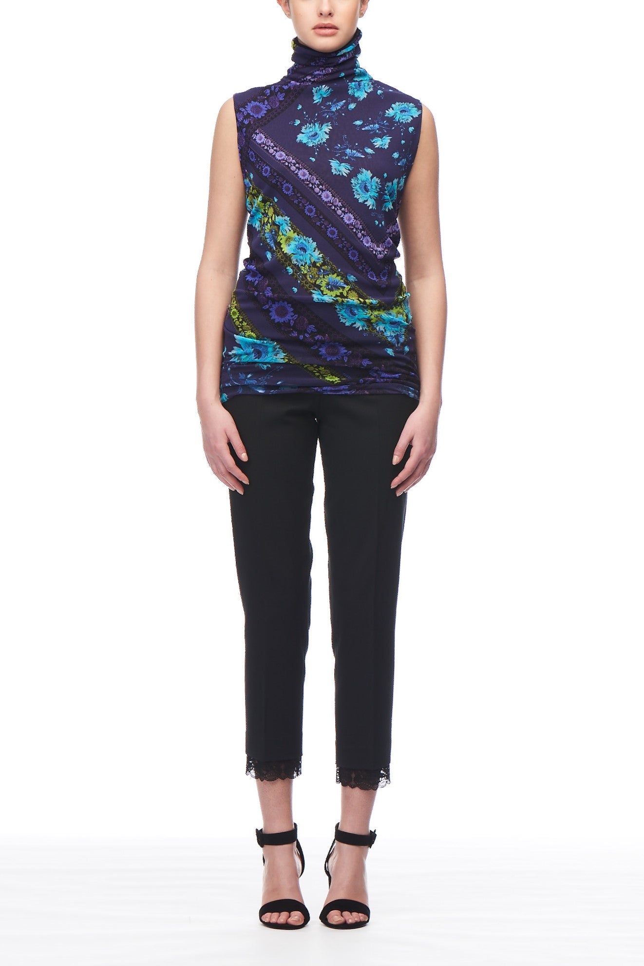 A person wearing the Fuzzi Woman Floral Mesh Tank by Fuzzi, featuring an ethereal floral print in shades of blue, purple, and green, paired with black cropped pants with lace trim at the hem. They complete the look with black strappy high heels.