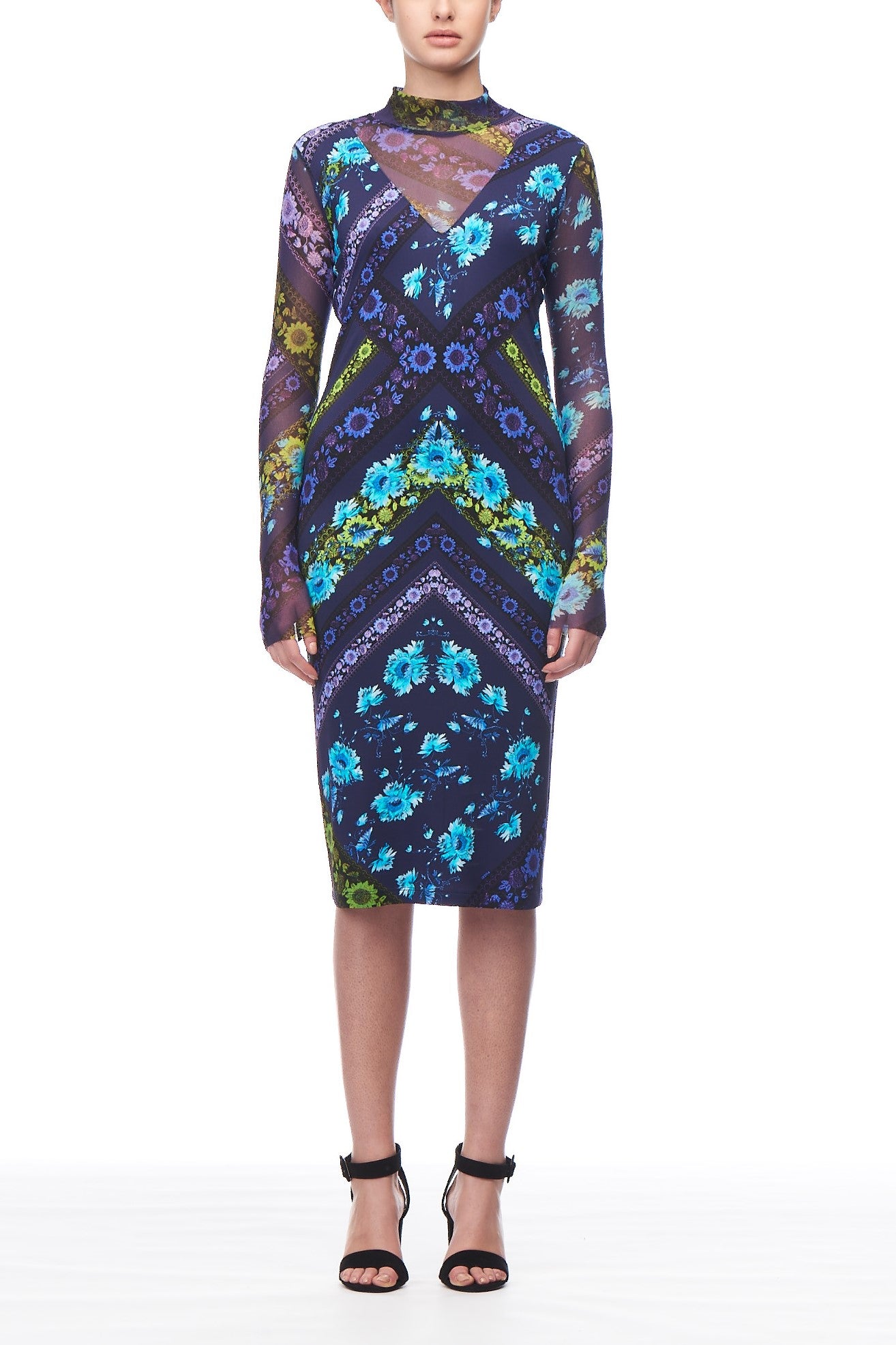 Dressed in the Fuzzi Woman Intricate Floral Dress, a person stands against a white background, showcasing bohemian-inspired elegance. The knee-length dress features long sleeves and a vibrant geometric floral pattern in shades of blue, purple, and green. Their look is completed with black high-heeled sandals.