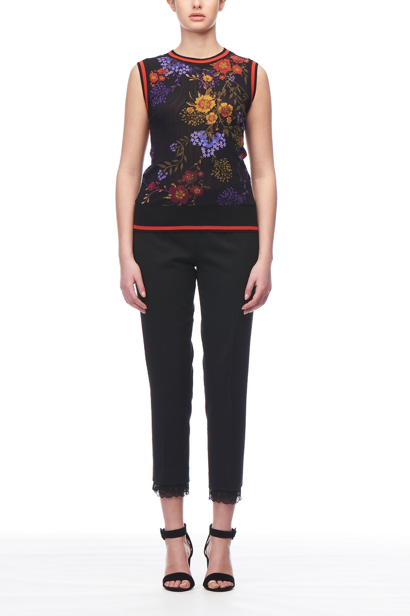 A person is wearing the Fuzzi Floral Mesh Tank, a sleeveless mesh top by Fuzzi adorned with vibrant floral patterns. They have paired it with black cropped pants featuring lace trim at the hem and are sporting black strappy heels, standing against a plain white background.