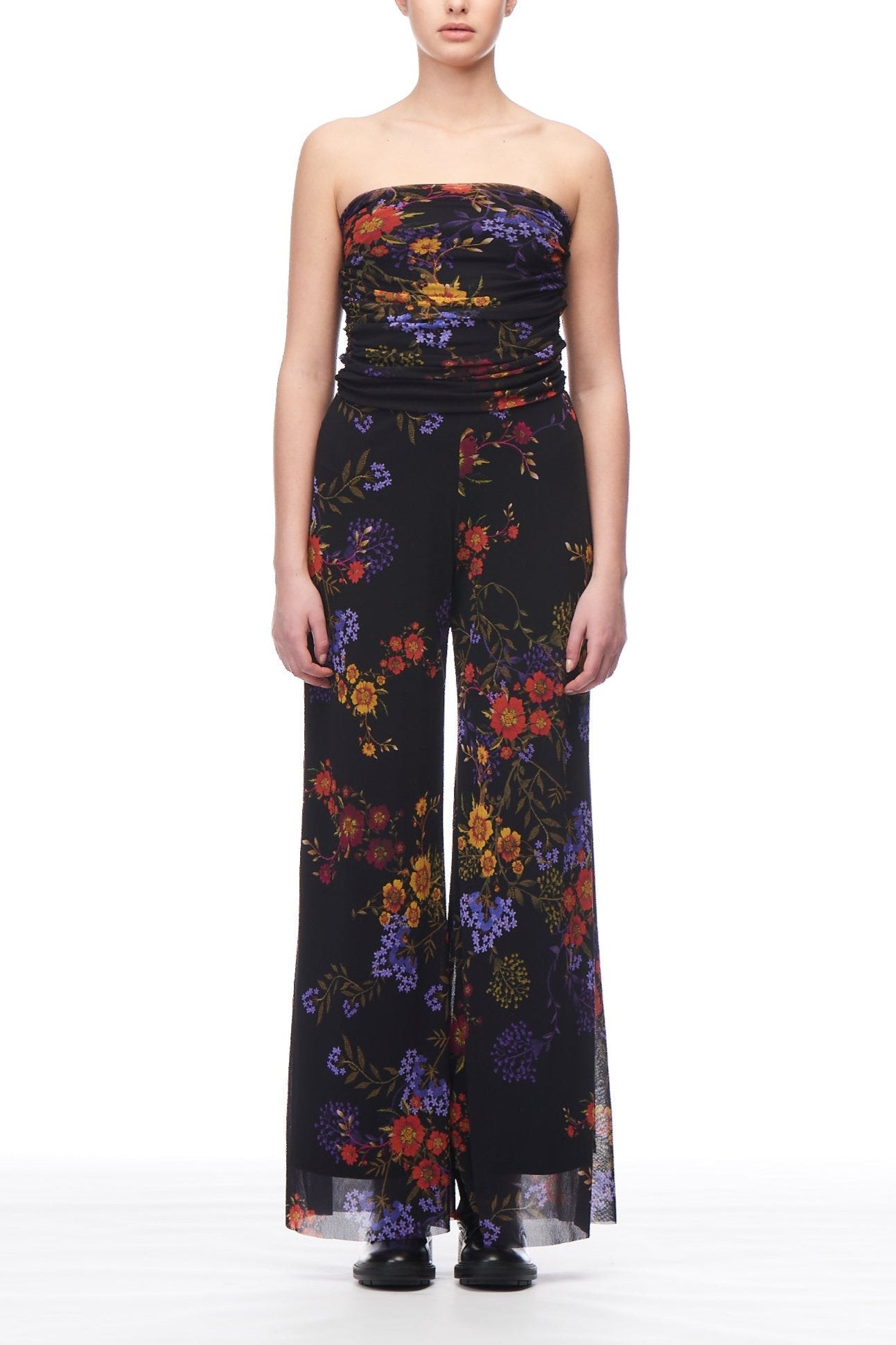 Against a white background, a person is seen wearing the Fuzzi Woman Floral Fantasy Jumpsuit by Fuzzi. This bohemian piece is adorned with colorful floral patterns in shades of red, orange, yellow, and purple. The strapless jumpsuit features wide-leg pants and is paired with black shoes to complete the look.