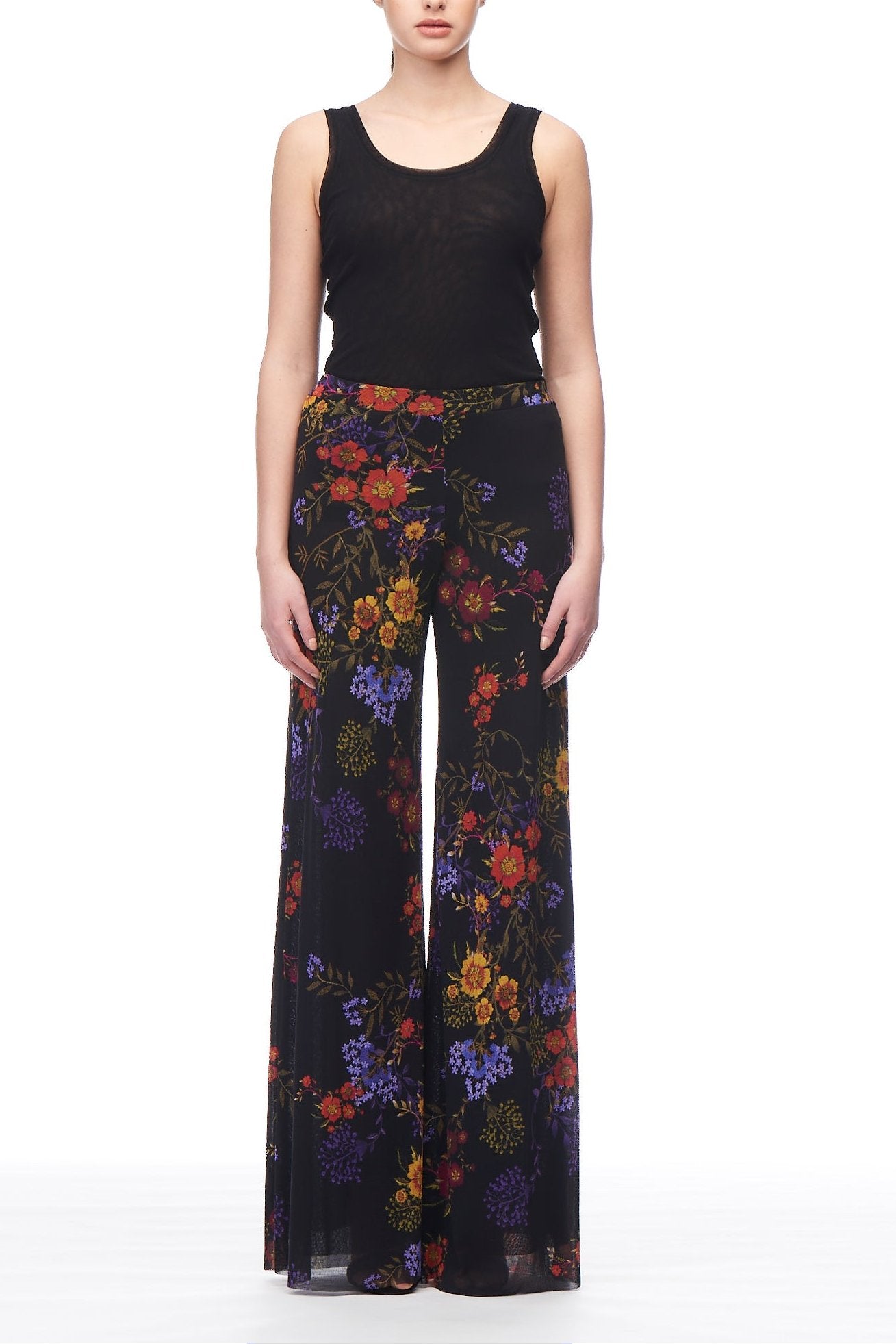 A woman poses against a white backdrop, elegantly showcasing the Fuzzi Floral Fantasy Trousers, which feature a colorful floral pattern and are paired with a black sleeveless top to enhance her silhouette.