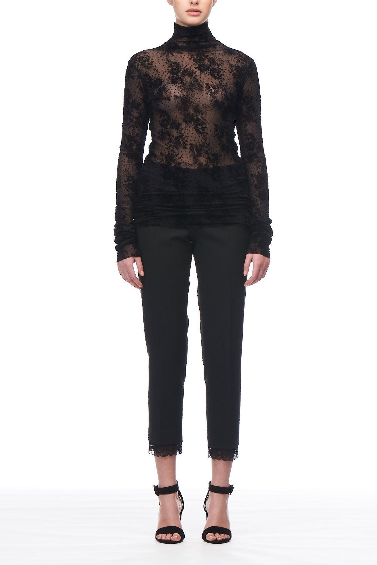 In front of a white background, a person models the Fuzzi Woman Mesh Top from Fuzzi, paired with black pants featuring lace trim. The ensemble is completed with sleek black high-heeled sandals, and their hair is pulled back neatly to highlight the outfit's alluring blend of textures and mesh fabric details.