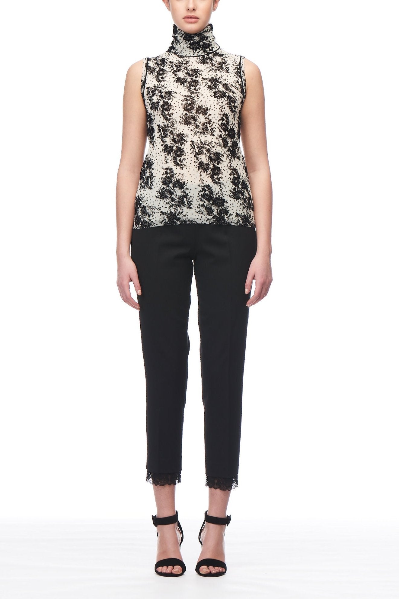 A person stands against a white background wearing the Fuzzi Woman Elegant Floral Turtleneck, featuring a black and white pattern in mesh fabric. They pair it with black cropped pants and black high-heeled sandals, while their hair is pulled back neatly.