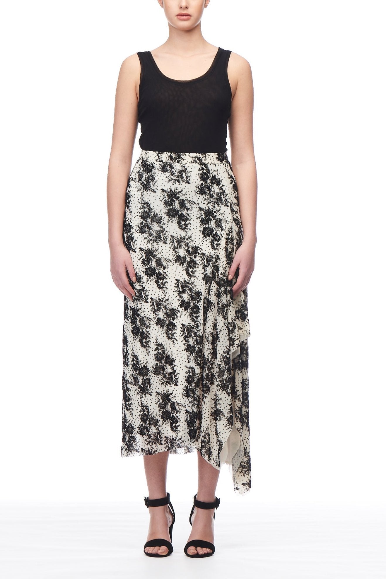 A woman stands against a white background, wearing a sleeveless black top paired with the Fuzzi Woman Asymmetrical Skirt, embellished with floral embroidery. She finishes her look with black high heels, and her hair is neatly pulled back.