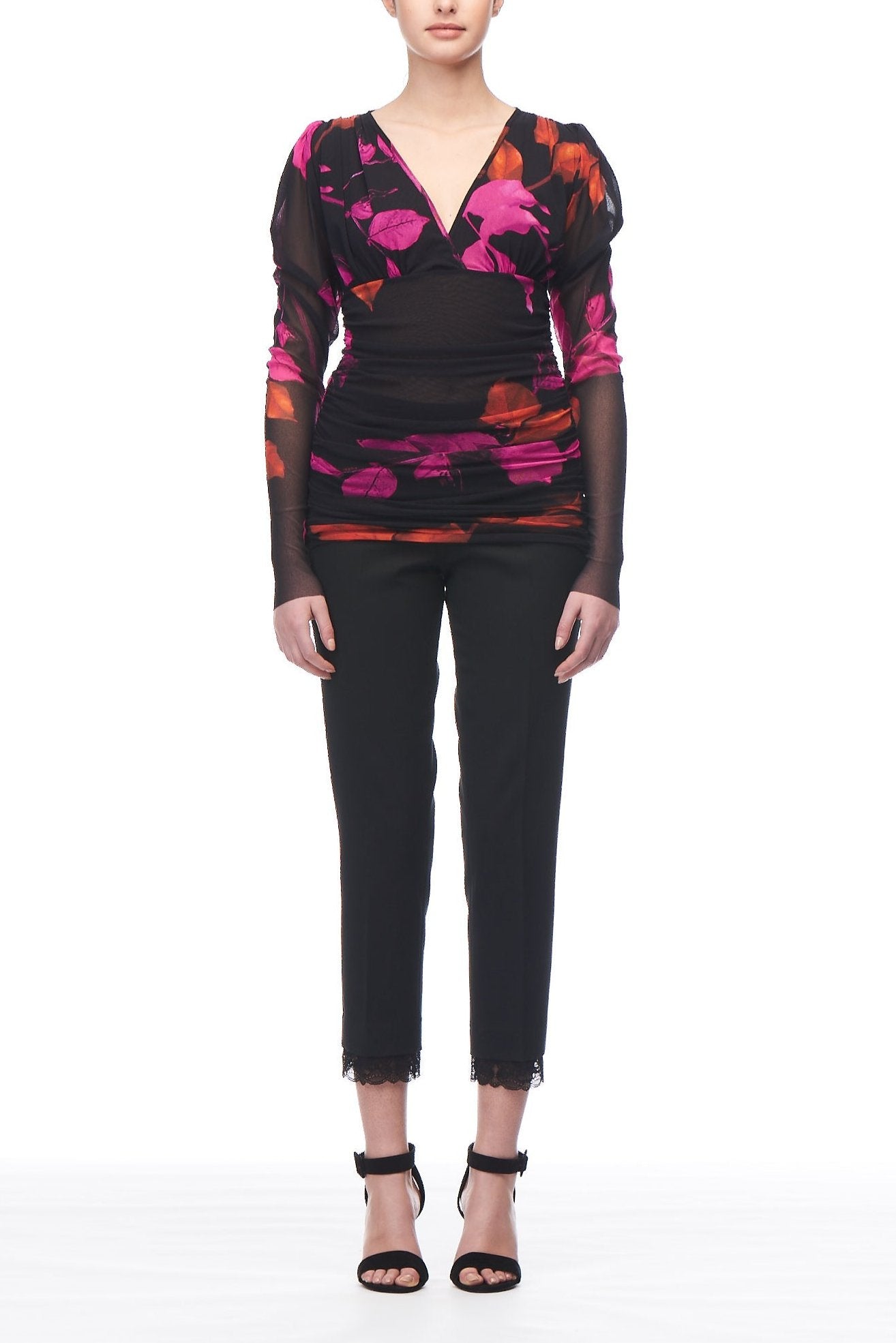 A person wearing the Fuzzi Woman Boho Chic Mesh Top, featuring a colorful floral print and long sleeves, paired with black slim-fit pants, stands against a white background. They have short hair and are wearing black high-heeled sandals.