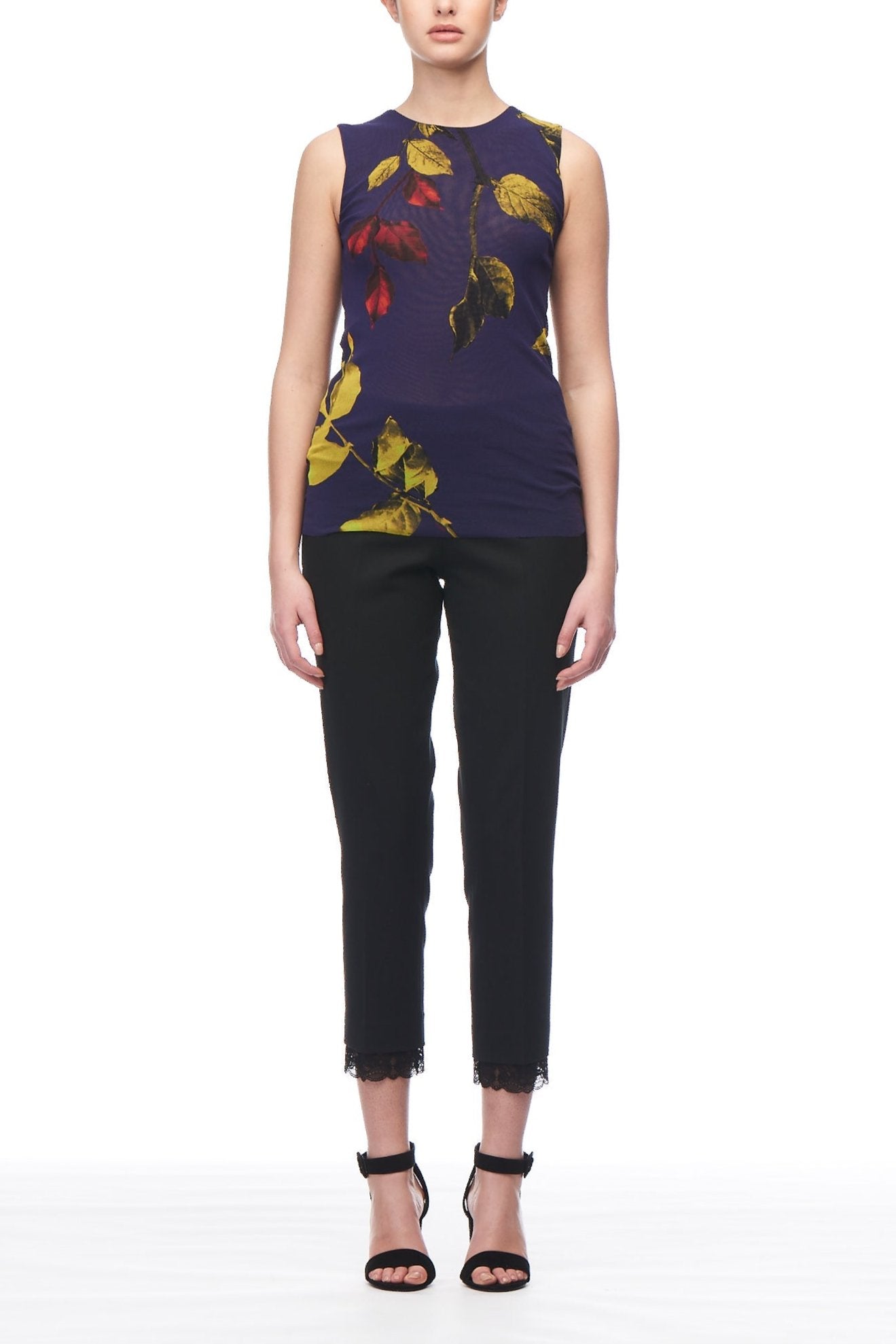 A person stands facing forward against a white background, wearing the Fuzzi Woman Sleeveless Mesh Top by Fuzzi with a Bohemian chic floral print, paired with black cropped pants and black high-heeled sandals. Their hair is styled back neatly.