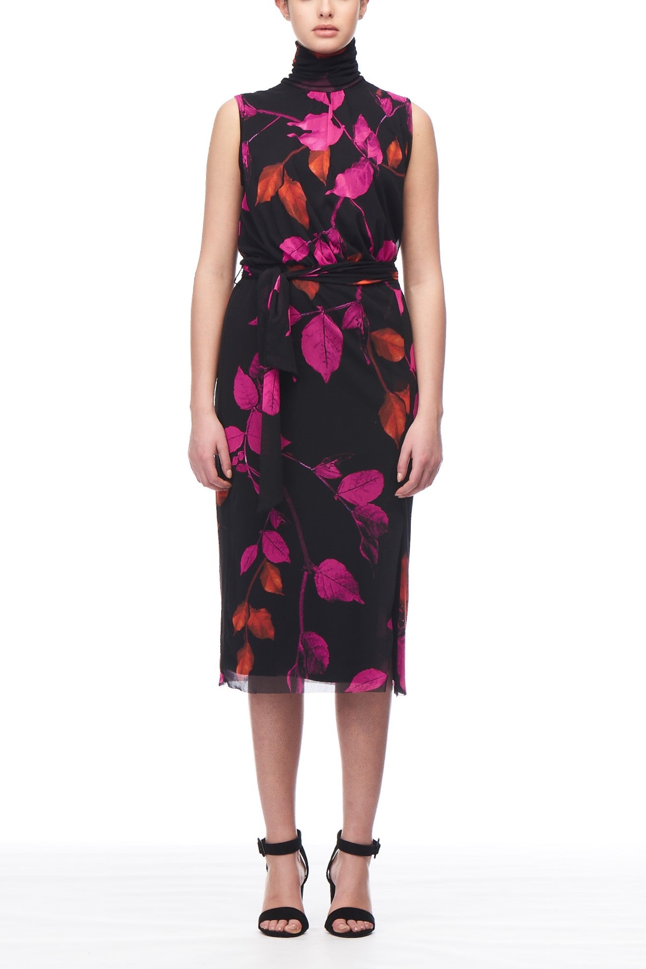 A person wears the Fuzzi Woman Floral Turtleneck Dress, a sleeveless black dress by Fuzzi featuring a vivid pink and red leaf pattern. This knee-length piece showcases an asymmetrical hemline and includes a matching waist tie. The ensemble is completed with black high-heeled sandals.