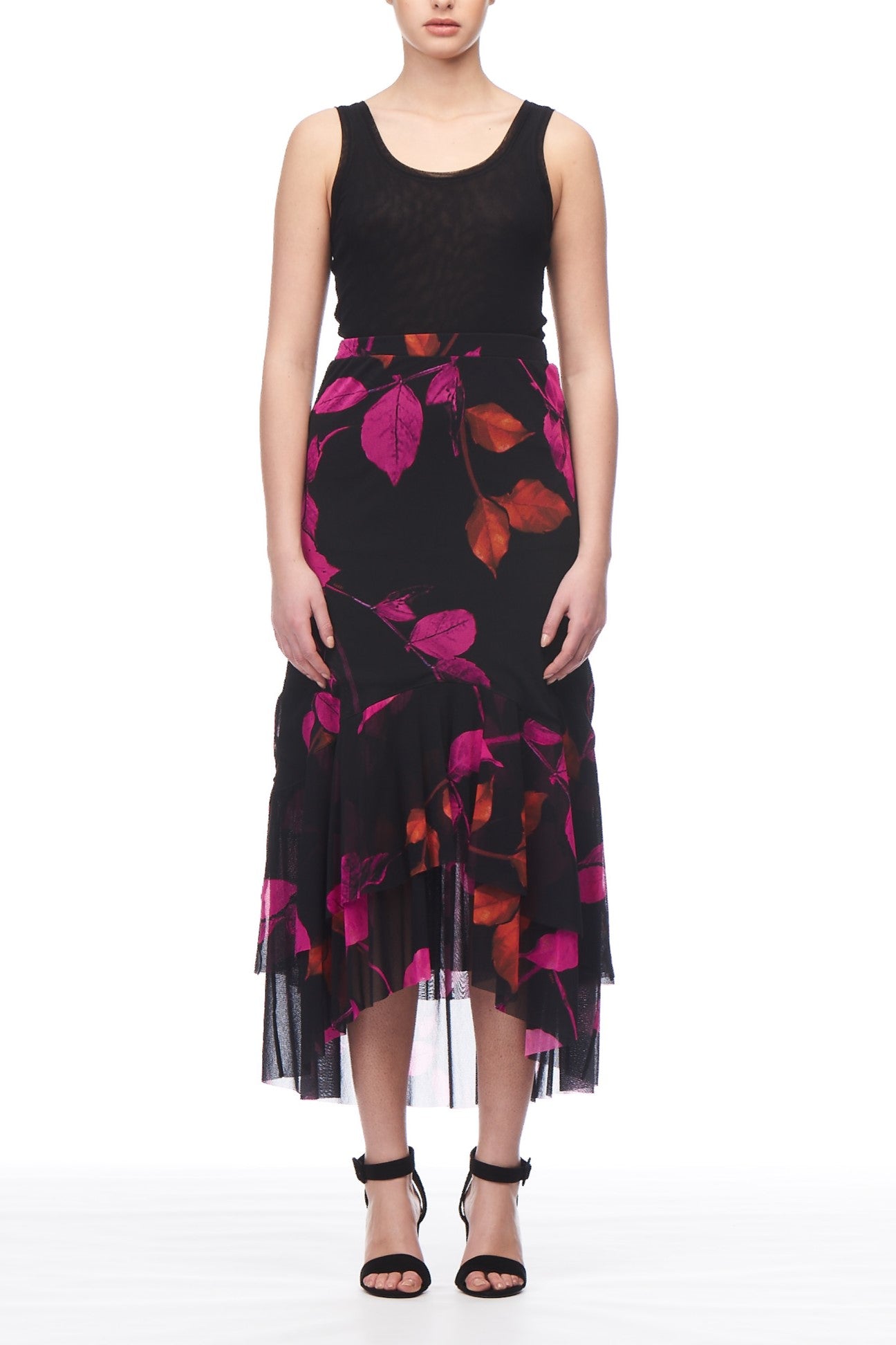 A person dressed in a sleeveless black top and the Fuzzi Woman Floral Midi Skirt by Fuzzi, featuring vibrant pink and orange floral patterns with an asymmetrical hemline. Standing with hands at their sides, they completed the look with black open-toed heels, exuding a feminine vibe against the white background.