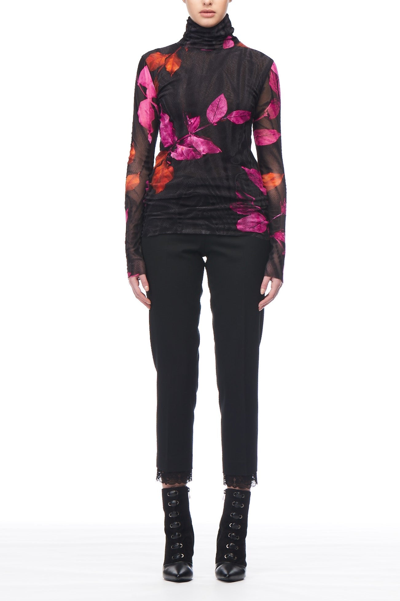 Wearing the Fuzzi Woman Floral Mesh Neck Top, adorned with a sheer black base and vibrant pink and orange floral design, a person exudes charm. They pair it effortlessly with black pants and vintage lace-up ankle boots, standing against a pristine white background.