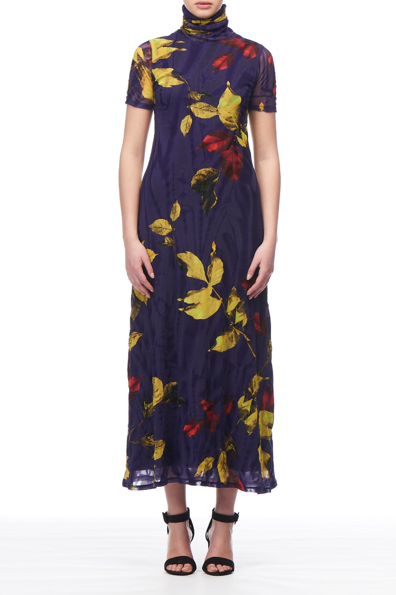 A person adorned in the Fuzzi Woman Floral Mesh Midi Dress, featuring a long purple midi silhouette with yellow and red floral patterns, stands against a white background. Perfect for a special occasion, this Fuzzi dress showcases short sleeves and a high neckline. The ensemble is completed with black high-heeled sandals.