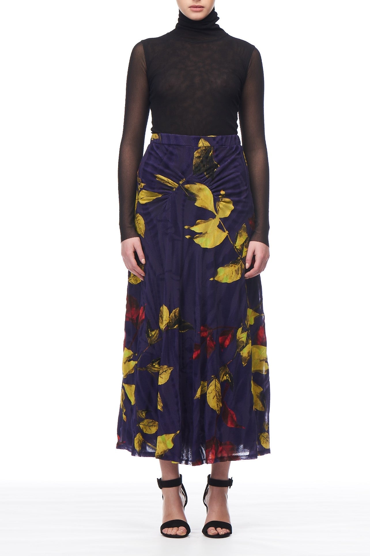 A person wears a black sheer turtleneck top paired with the Fuzzi Woman Midi Skirt by Fuzzi, featuring vibrant yellow and red blossoms on a dark blue background. Black high-heeled sandals complete the ensemble against the plain white backdrop.