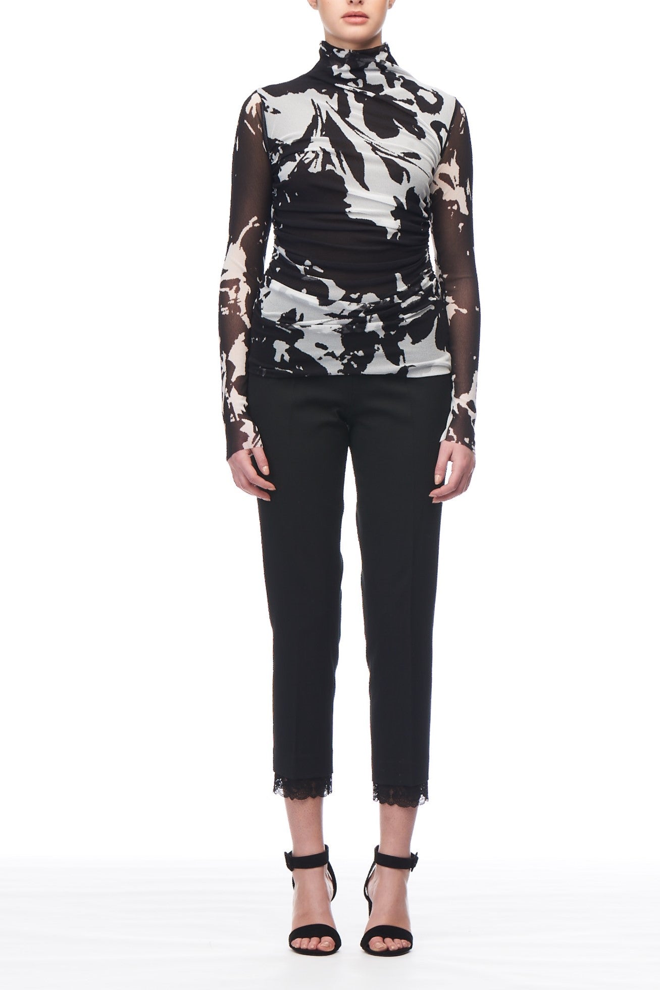 The ensemble showcases a Fuzzi Woman Turtleneck Top with an abstract print, featuring sheer mesh sleeves for a striking statement. Complemented by black cropped pants adorned with lace trim and paired with chic black high-heeled sandals, this outfit effortlessly combines style and boldness.