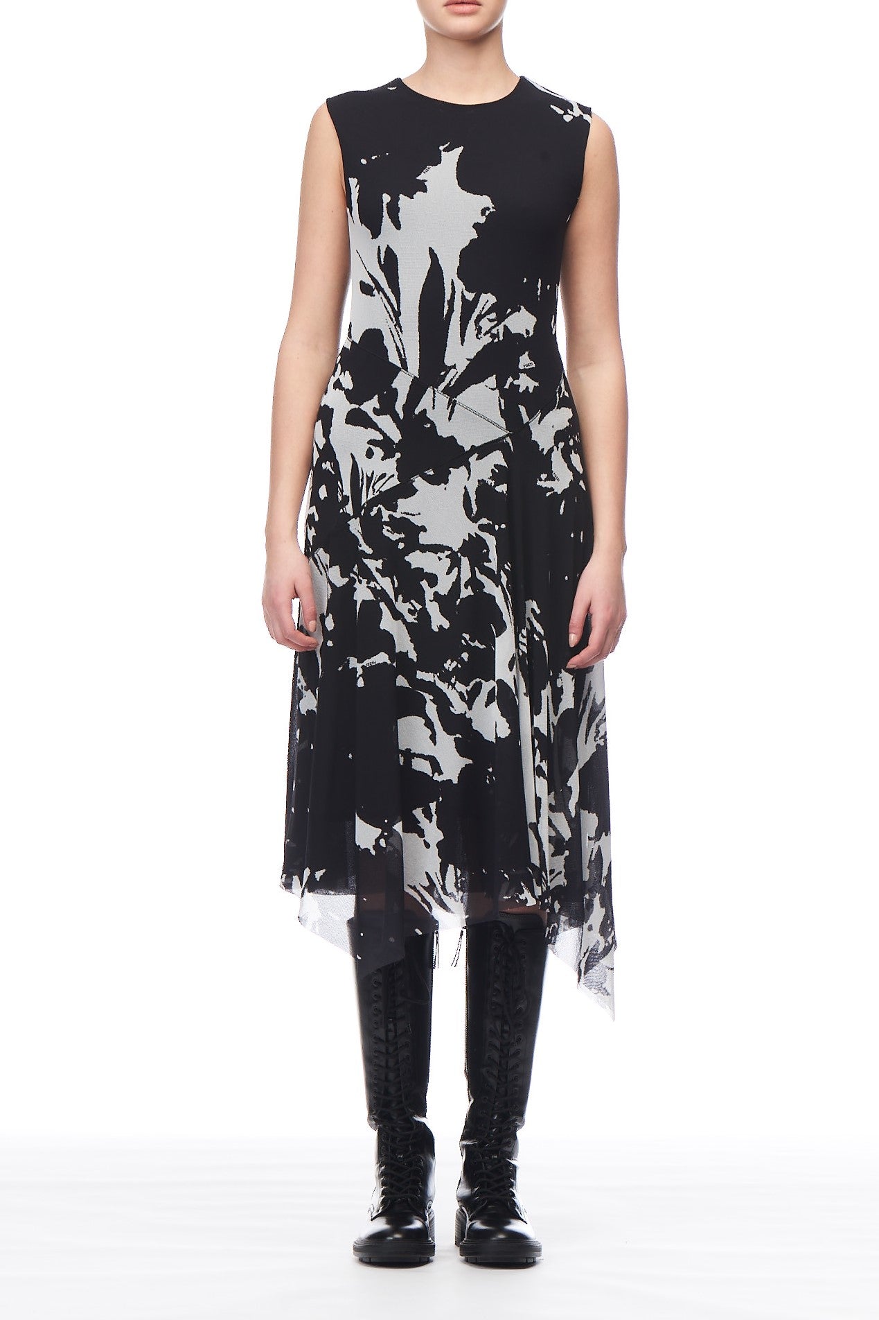 A person wearing the Fuzzi Woman Black and White Abstract Print Dress, a chic sleeveless piece from Fuzzi featuring an abstract floral pattern. The knee-length uneven hem adds elegance, stylishly paired with black lace-up boots. The plain white backdrop accentuates the sophisticated ensemble.