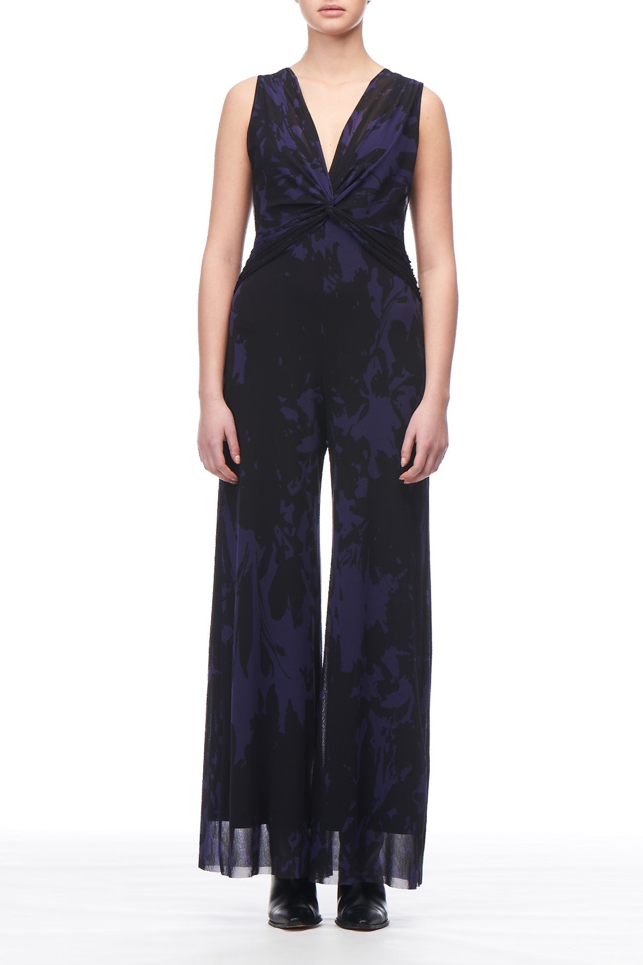 The individual stands facing forward, dressed in the Fuzzi Woman Sleeveless Jumpsuit by Fuzzi, which features a deep V-neck and a twist front detail. The fabric showcases a dark, swirling floral print. They are also wearing black shoes paired with stylish wide-leg pants against a plain white background.