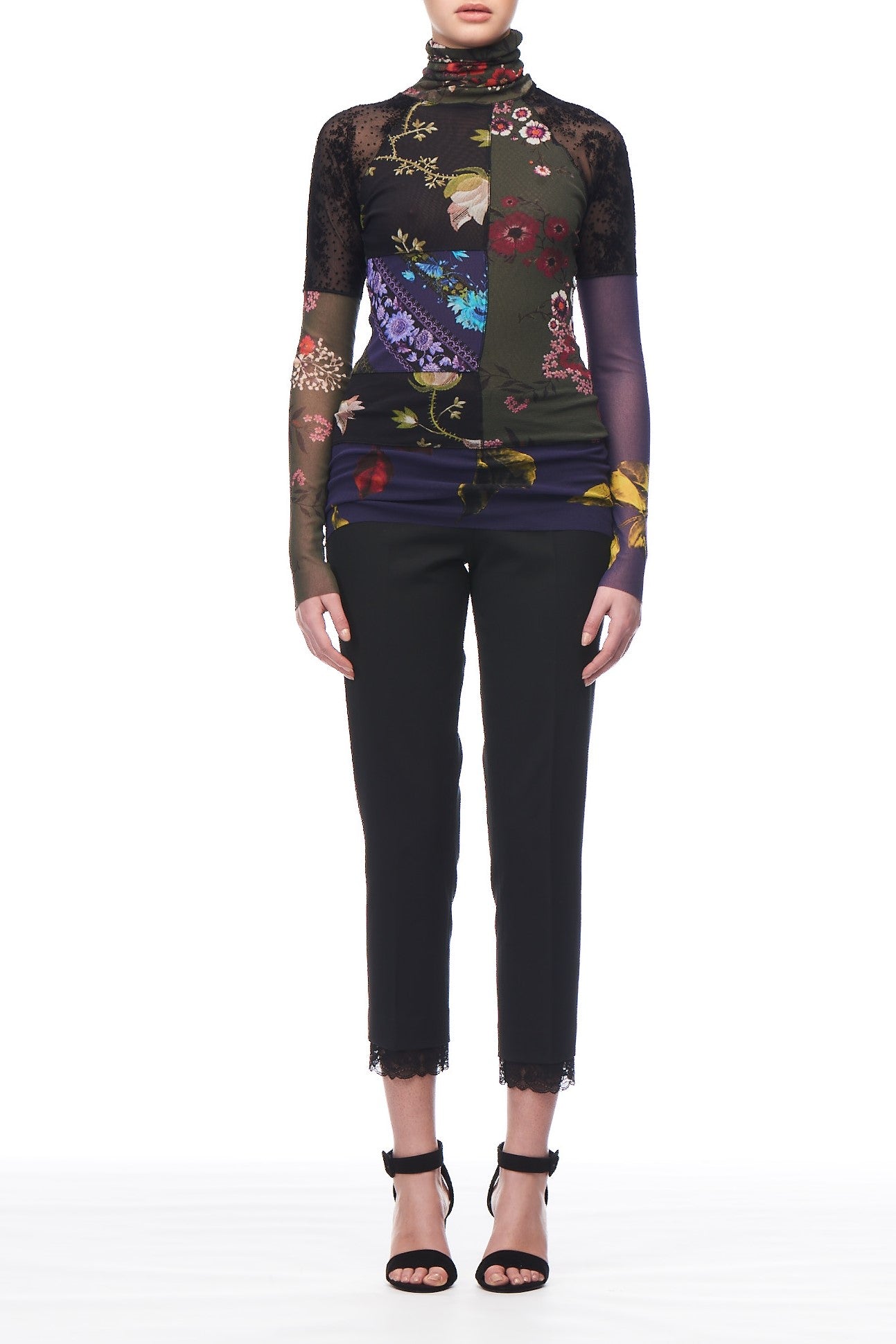 A person showcases the Fuzzi Woman Ethereal Floral Top, a high-neck, long-sleeve design featuring a vibrant abstract floral pattern and black semi-transparent panels. They pair it elegantly with black cropped pants and high-heeled shoes adorned with ankle straps, all set against a plain white background.