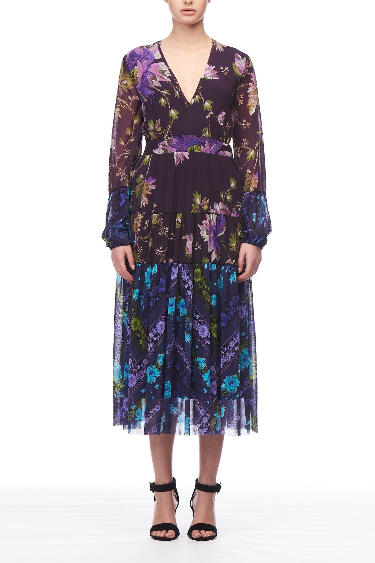 A person wearing the Fuzzi Woman Enchanted Floral Dress, a vintage piece with long sleeves and a floral pattern in shades of purple, blue, and green. This flowing silhouette from Fuzzi features a V-neckline and a tiered skirt, perfectly complemented by black strappy heels against the plain white background.