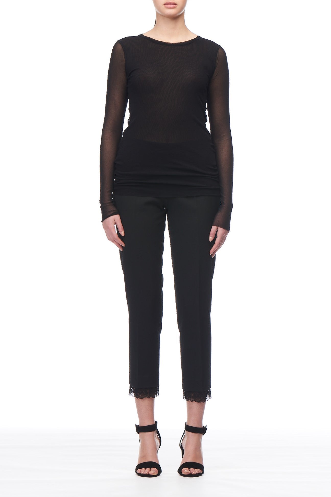 A person exudes sheer elegance in a Fuzzi Woman Delicate Mesh Top paired with black pants, standing against a white background. The cropped pants feature lace at the hem and effortlessly pair with black high-heeled sandals.