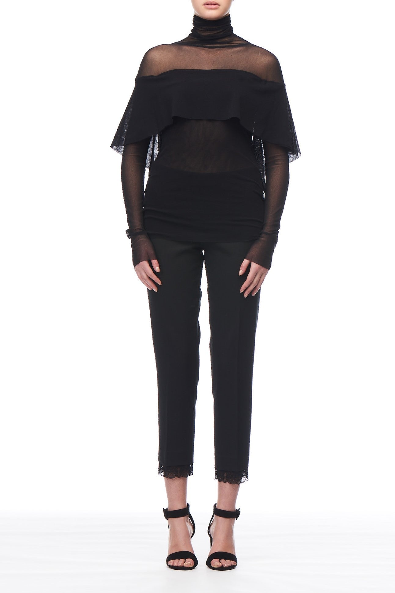 A person is wearing the Fuzzi Woman Black Mesh Top, featuring a sheer mesh fabric in black with a layered cape detail, over a black turtleneck. It's paired with fitted black pants and strappy high-heeled sandals against a white background.