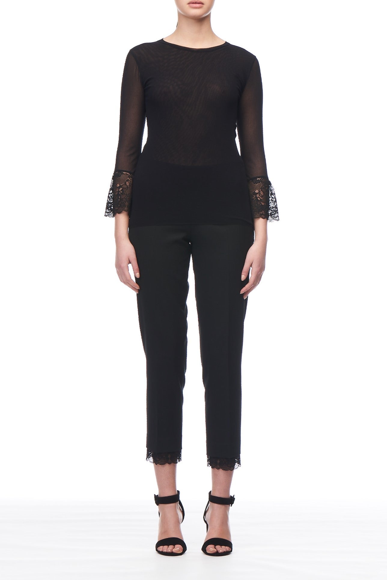 A person dressed in an elegant black ensemble, featuring the Fuzzi Woman Lace Cuffs Top—a sleek sheer mesh top with delicate lace sleeves. Complementing the look are black pants with vintage fashion lace trim at the hem and stylish black ankle-strap heels. The minimalist white background accentuates this sophisticated statement piece from the brand Fuzzi.