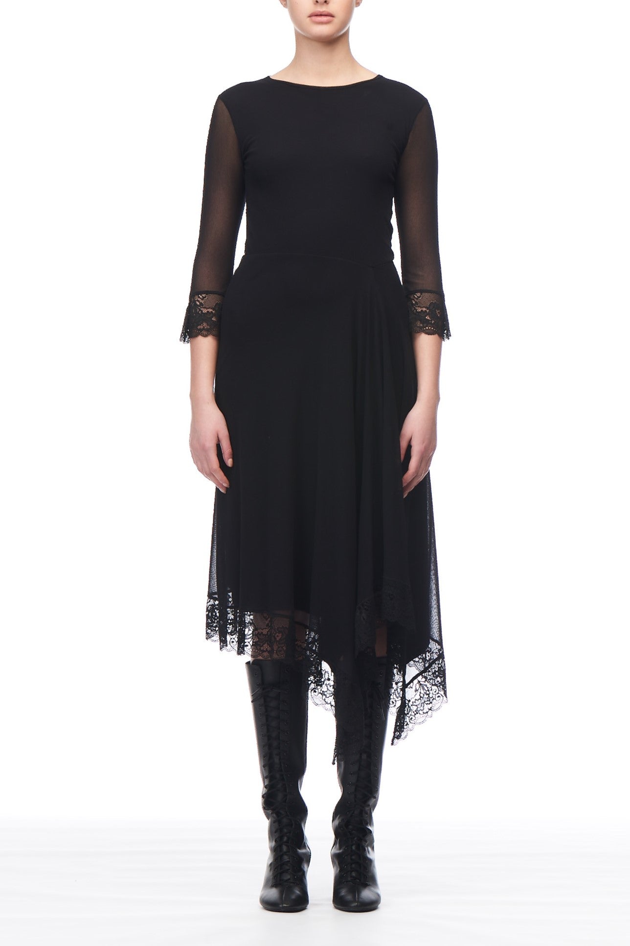 A person is dressed in a Fuzzi Woman Asymmetrical Lace Dress, featuring lace sleeves and hemline, a fitted bodice, and an asymmetrical skirt. They have paired the black vintage dress with knee-high black boots against a plain white background.