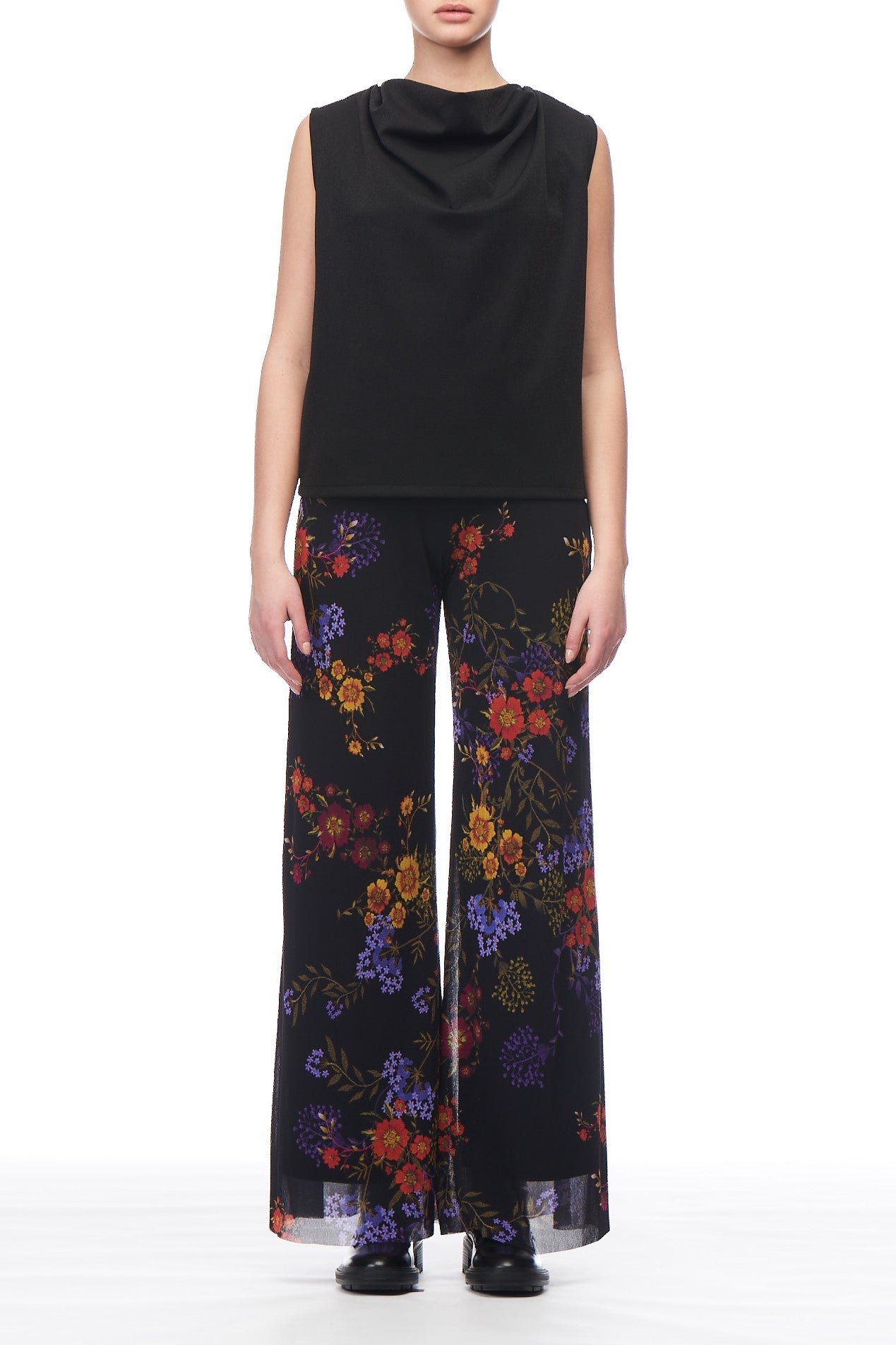 A person in a Fuzzi Woman Cowl Neck Sleeveless Top and wide-leg black pants with colorful floral patterns exudes a modern, stylish vibe against the white background. The ensemble, paired with sleek black shoes, subtly hints at vintage piece inspiration while maintaining its contemporary charm.