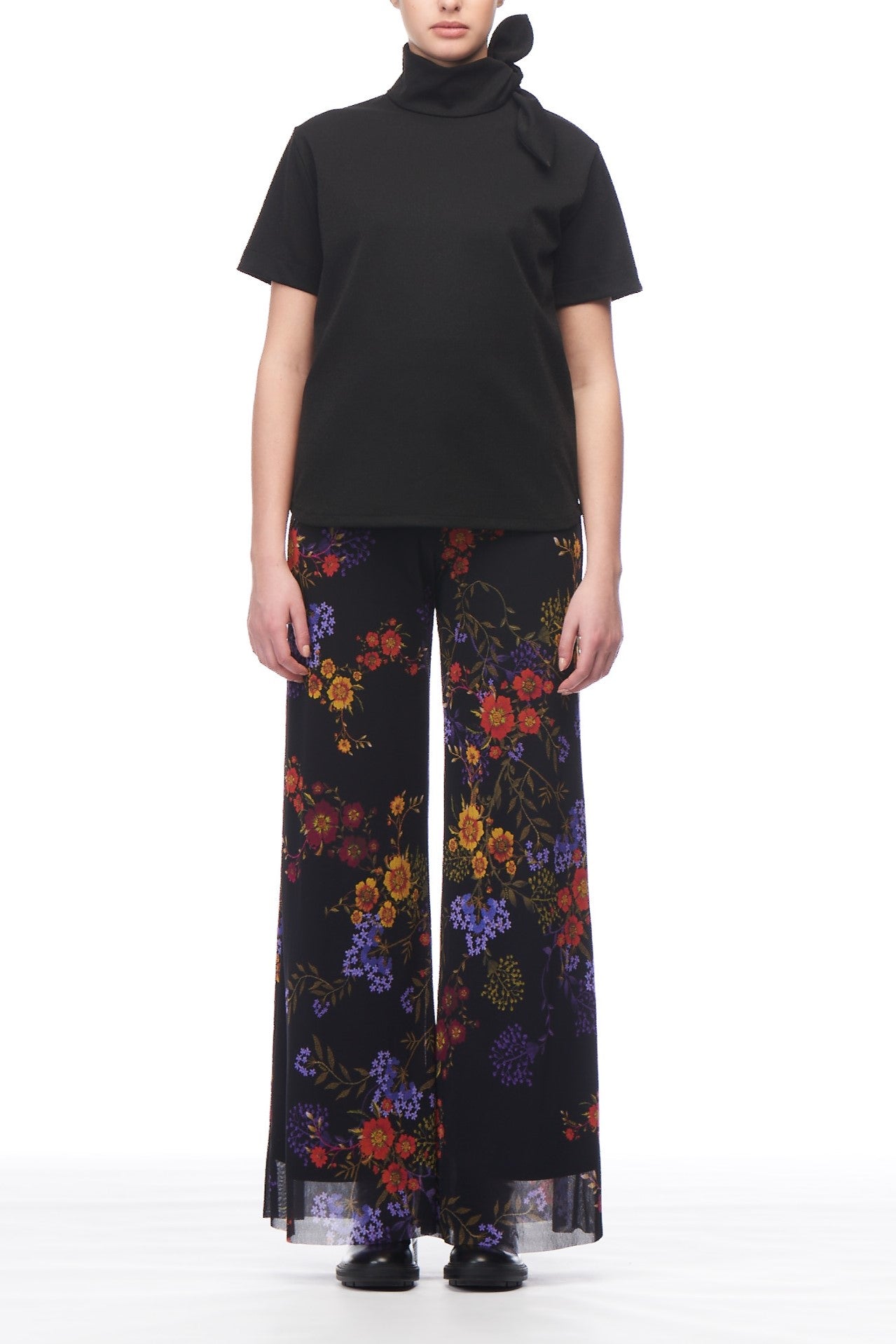 A person is wearing the Fuzzi Woman Ruffled High Neck T-Shirt, a black sleeveless top with bow detailing. It is paired with vibrant floral pattern-wide-leg pants, creating a vintage look that's completed by black shoes, all set against a plain white background.