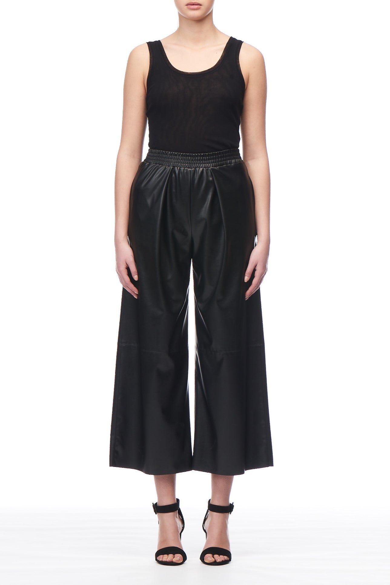 Against a simple white backdrop, a person is elegantly outfitted in a sleeveless black top paired with the Fuzzi Woman High-waisted Trousers by Fuzzi, boasting a wide-leg silhouette crafted from shiny material, along with stylish black strappy heels.