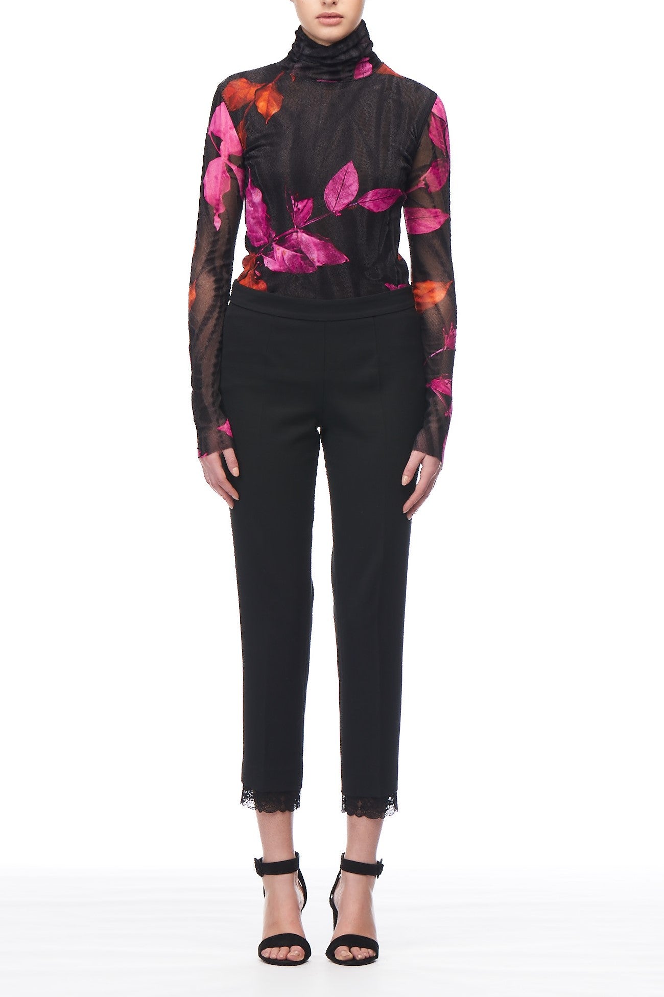 The individual is dressed in a sheer black top featuring vibrant pink leaf patterns and Fuzzi Woman Black Trousers from the brand Fuzzi, highlighted by vintage lace trim details. They complete the ensemble with black open-toe heels, all set against a plain white background.