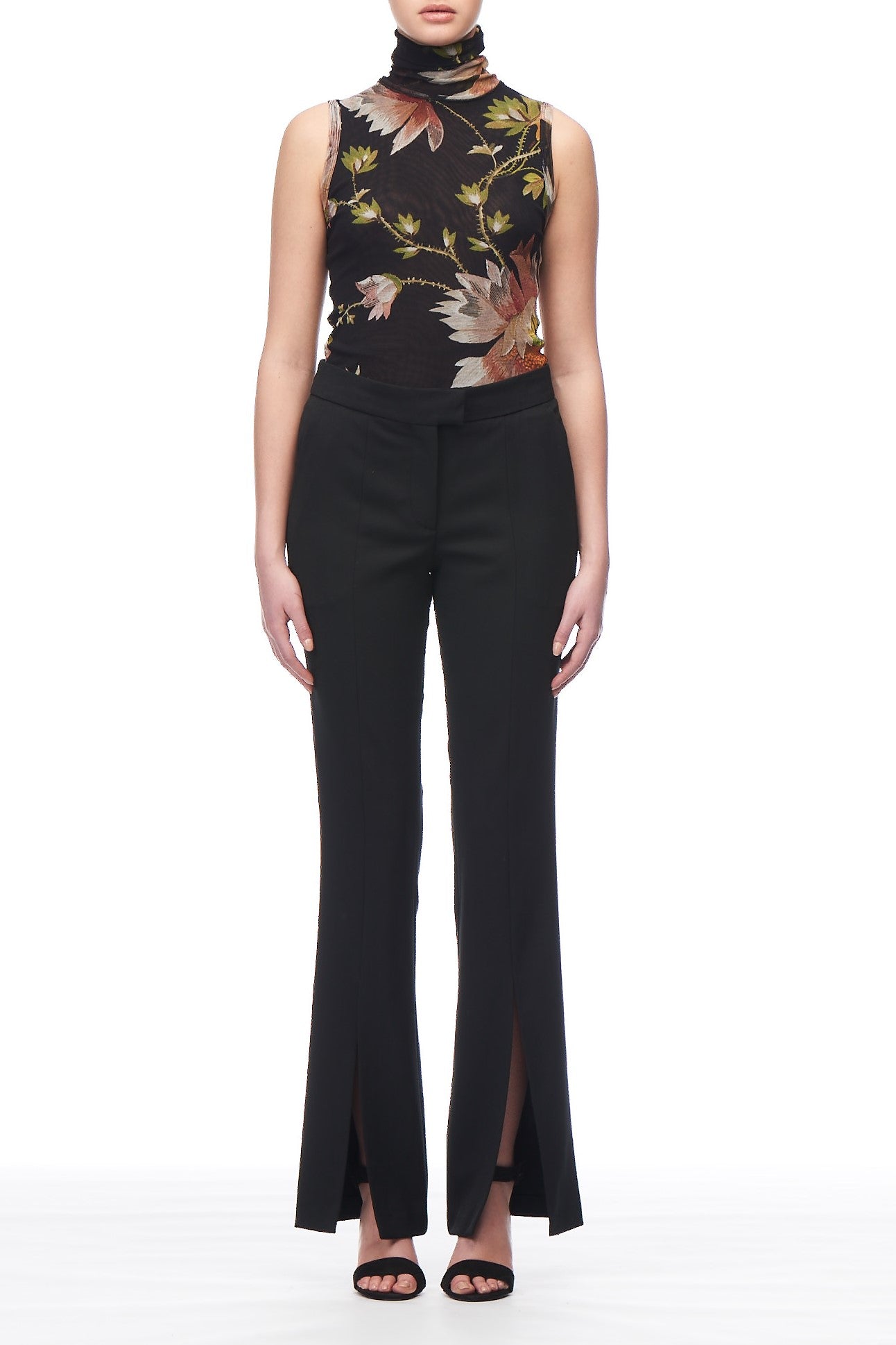 A person is wearing a floral-patterned high-neck sleeveless top with Fuzzi Woman Black Trousers by Fuzzi, which feature front slits. They complete the ensemble with open-toed shoes against a plain white background.