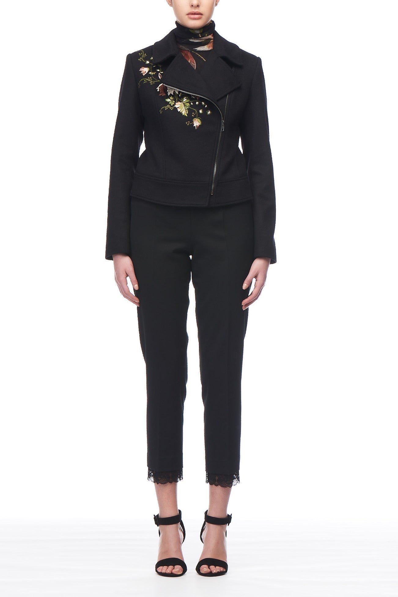 A person is elegantly dressed in a chic Fuzzi Woman Embroidered Jacket featuring stylish floral designs, coordinated with vintage black pants highlighted by delicate lace trim at the ankles, and complemented by sleek black high-heeled sandals. The setting is against a simple, pristine white backdrop.