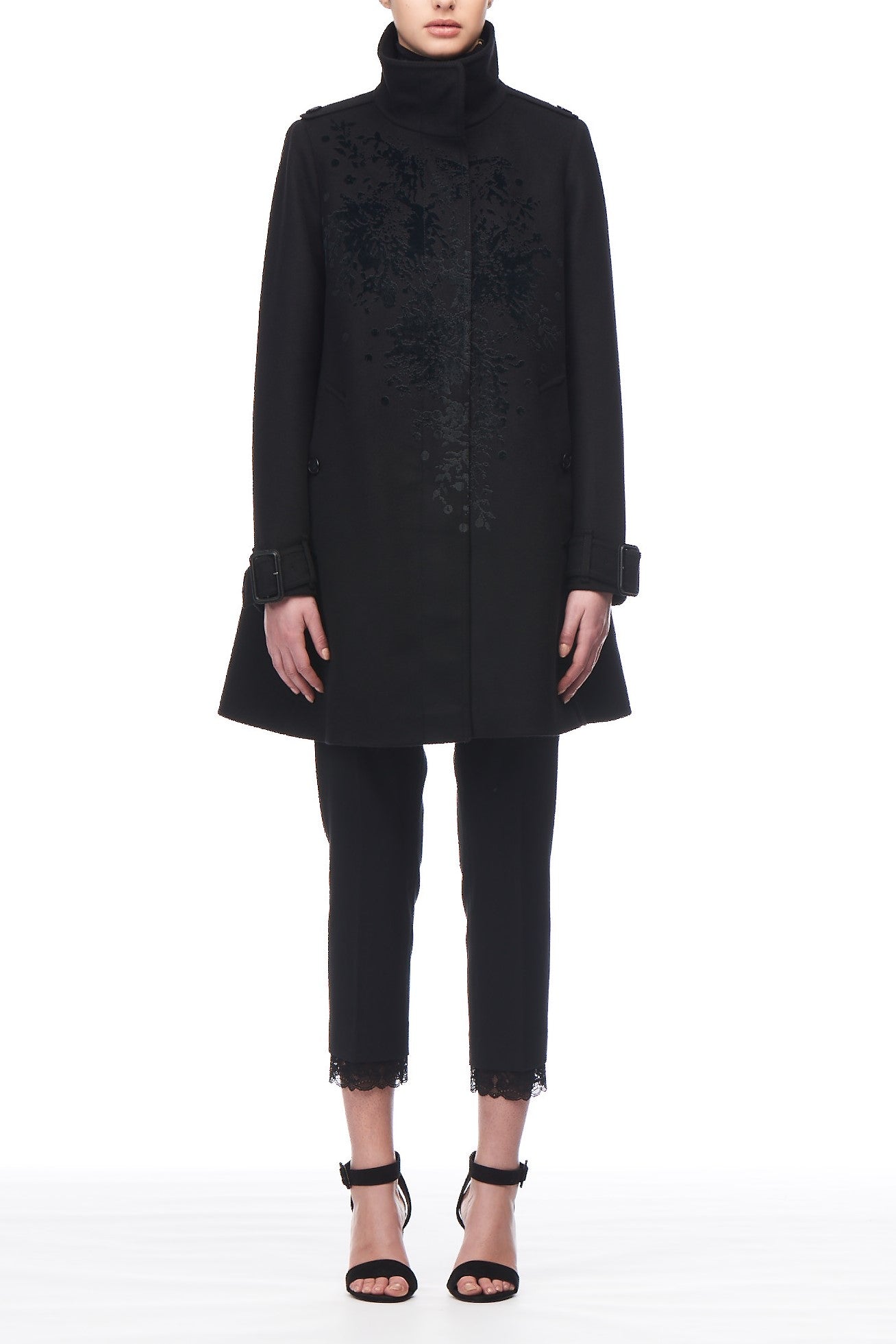 A person is modeling the Fuzzi Woman High Neck Coat, which features velvet embroidery, paired with cropped dark pants and strappy black heels against a plain white background.