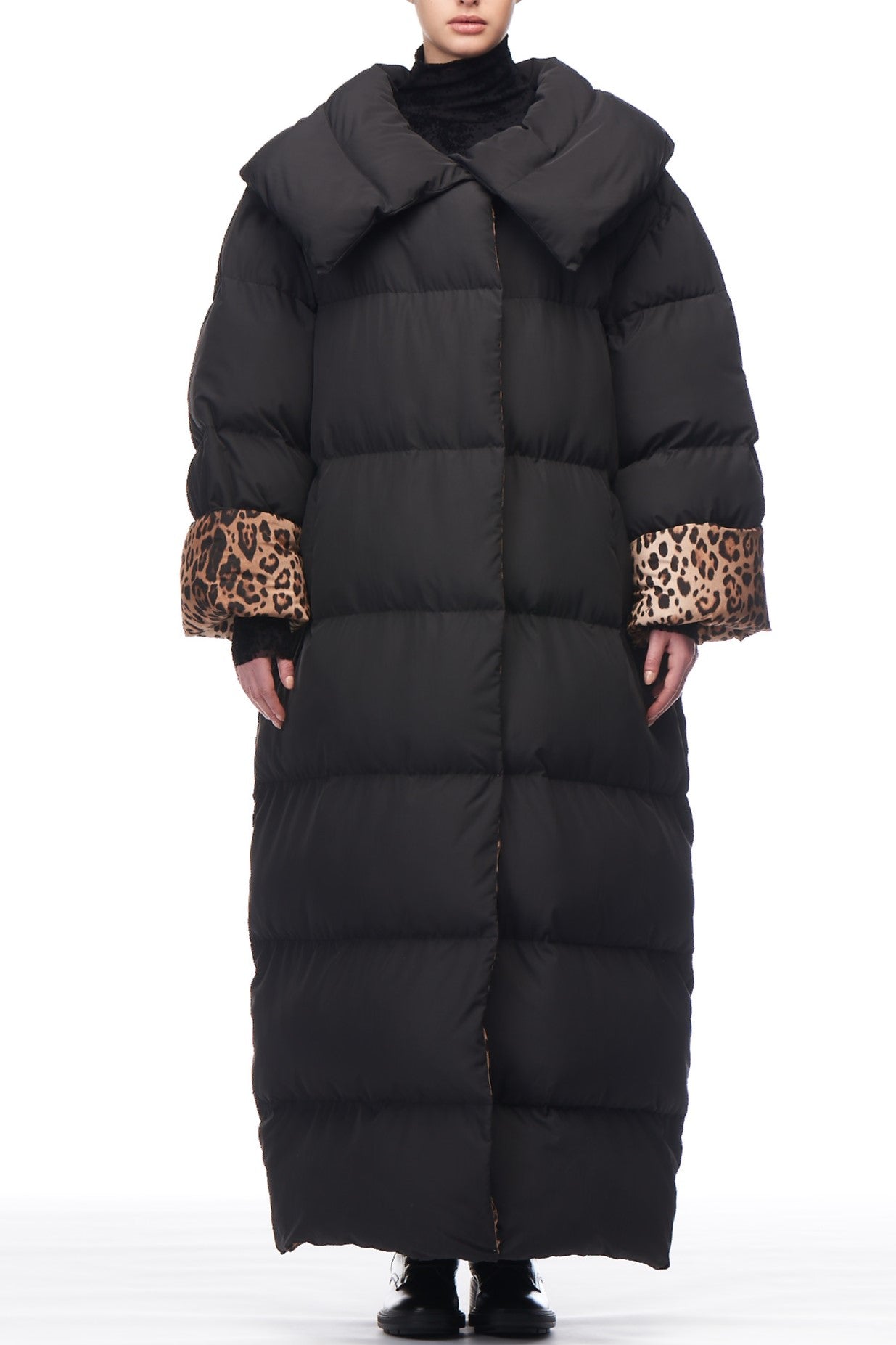 During the winter season, a person wears the Fuzzi Woman Leopard Trim Puffer Coat by Fuzzi, featuring a long black design with an eye-catching large collar and leopard print cuffs. The coat elegantly flows to the ankles, perfectly complementing black shoes against a crisp white background.