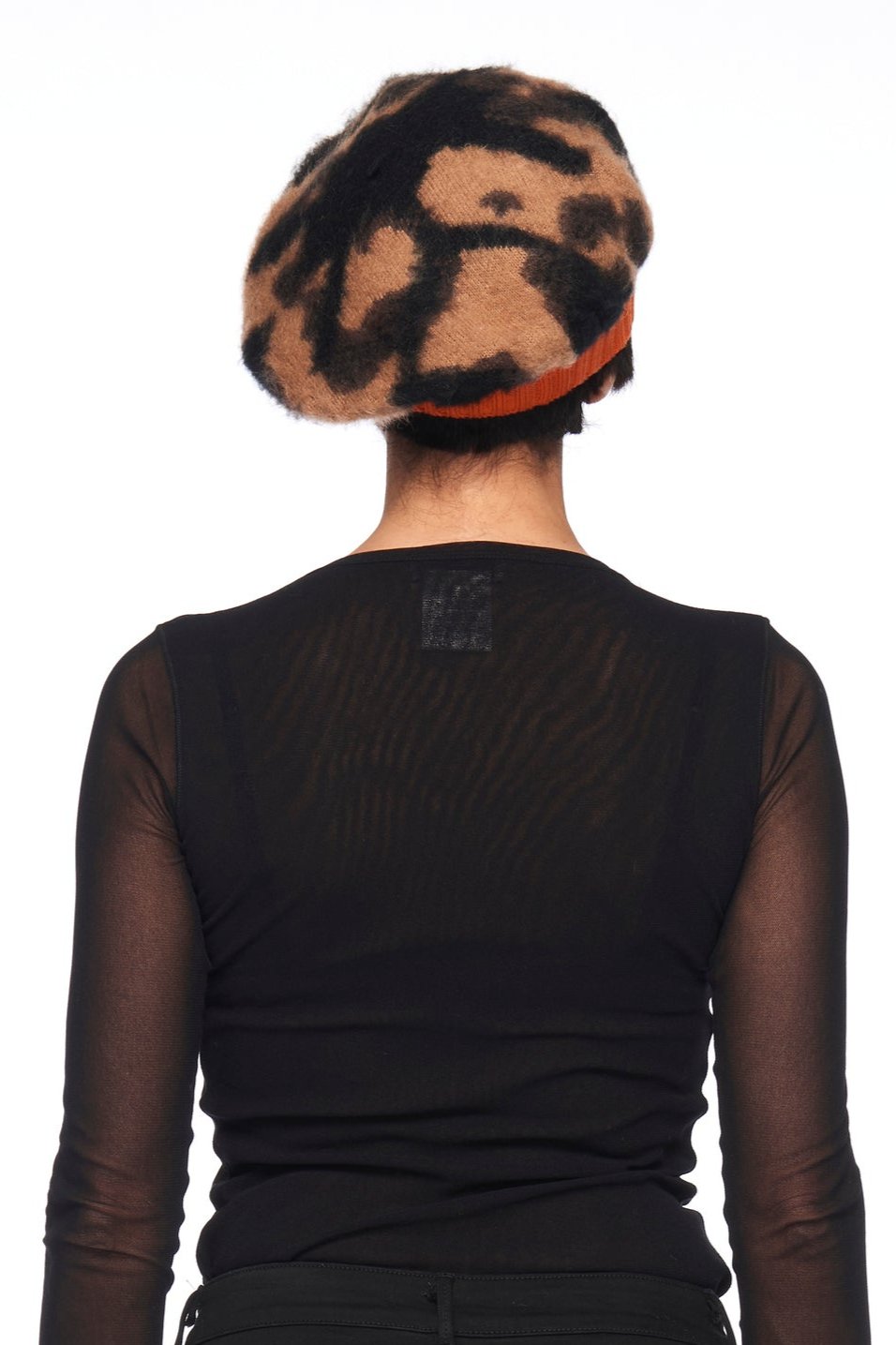 A person is facing away, wearing a sheer black top and the Fuzzi Woman Abstract Print Beanie with an orange band against a plain white background.