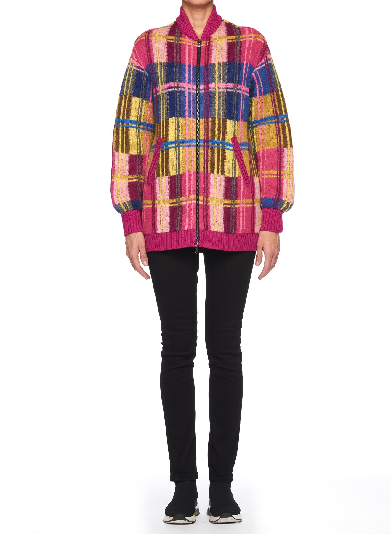 A person showcasing a vibrant, retro-inspired fashion piece—the Fuzzi Woman Bomber Jacket with a plaid pattern featuring shades of pink, yellow, blue, and black—paired with black pants and shoes. The oversized fit contributes to their stylish appearance as they pose against a white backdrop.