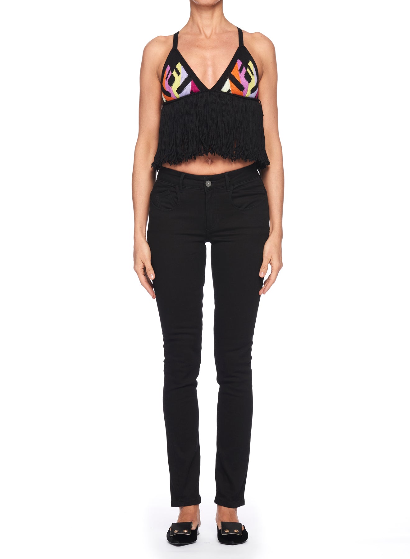 A woman poses against a white background, wearing the Fuzzi Woman Sleeveless Top featuring black fringes and colorful abstract patterns with a fitted silhouette, paired with black skinny jeans. Her look is completed with sleek black shoes as her arms rest calmly at her sides.