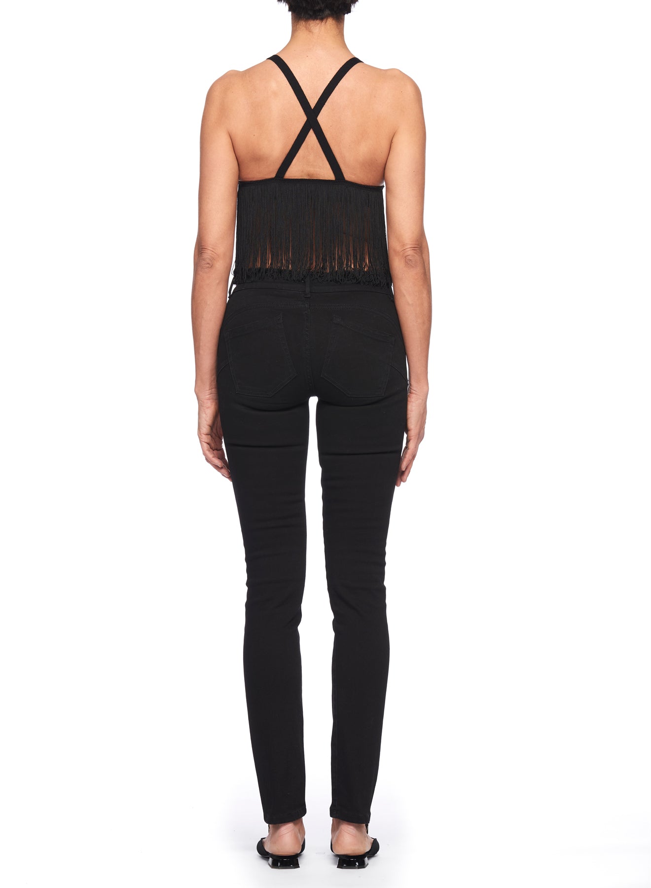 A person wearing the Fuzzi Woman Fringed Top in boho chic black pairs it with black jeans as they stand facing away from the camera. Their short hair complements their look, completed with a pair of black open-toe shoes, all against a plain white background.