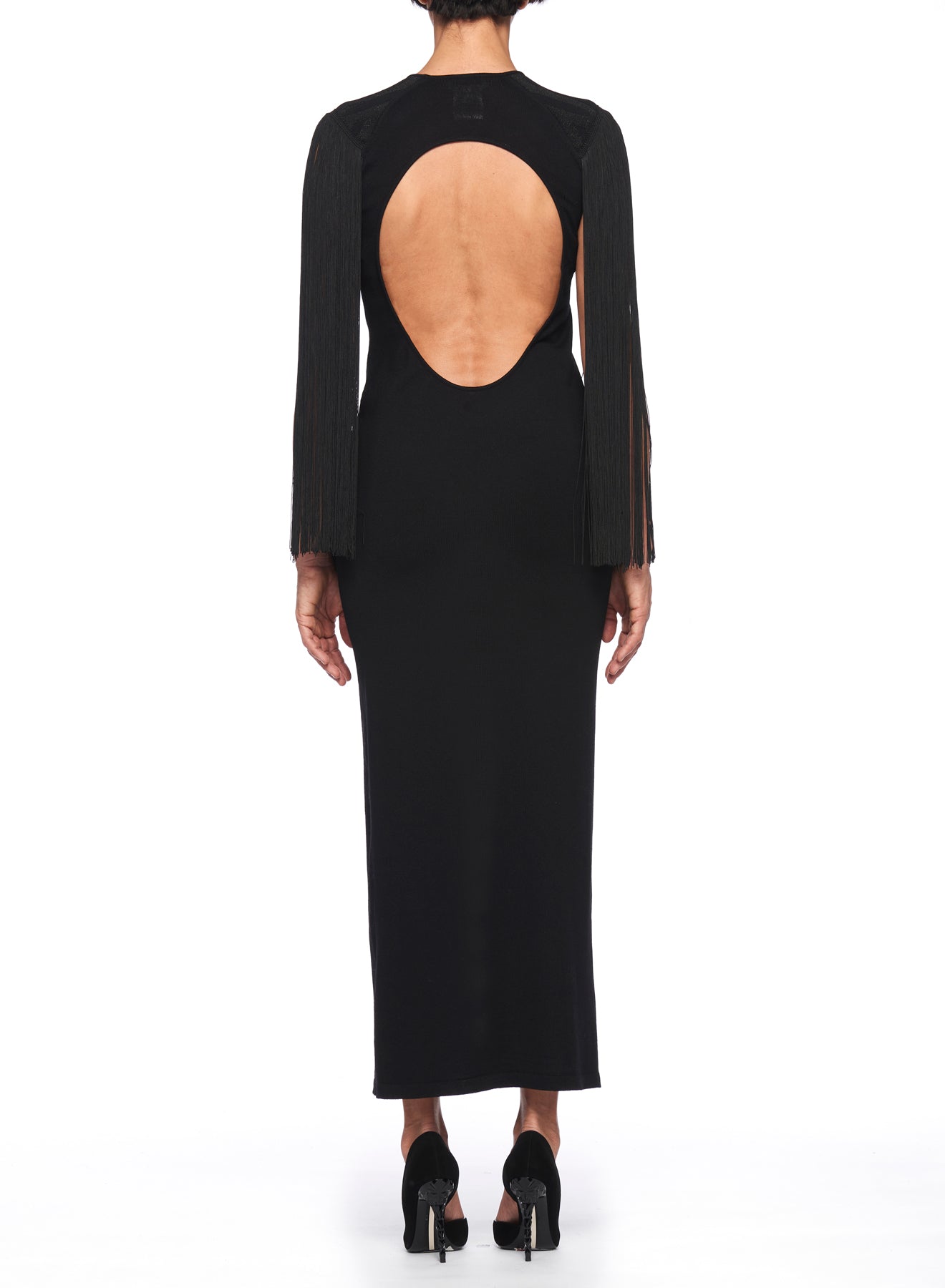 A person wearing the stunning Fuzzi Woman Backless Dress, characterized by its high neck and long, fringed sleeves, stands against a white background. This form-fitting black ensemble is perfectly paired with sleek black high-heeled shoes.