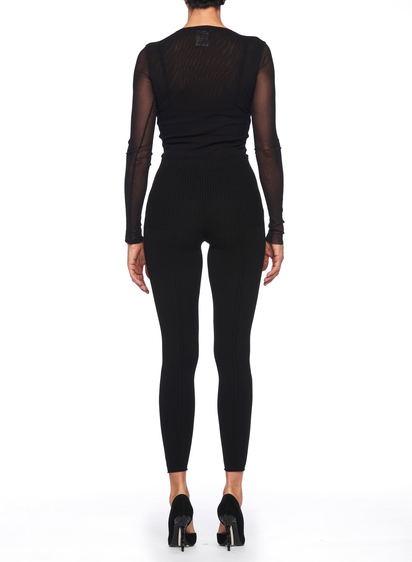 A woman stands with her back to the camera, highlighting a sheer black long-sleeve top paired with Fuzzi Woman's Sleek Ribbed Leggings, which feature a high-waisted design. Accentuated by black high-heeled shoes, this outfit exudes timeless elegance against the white background.