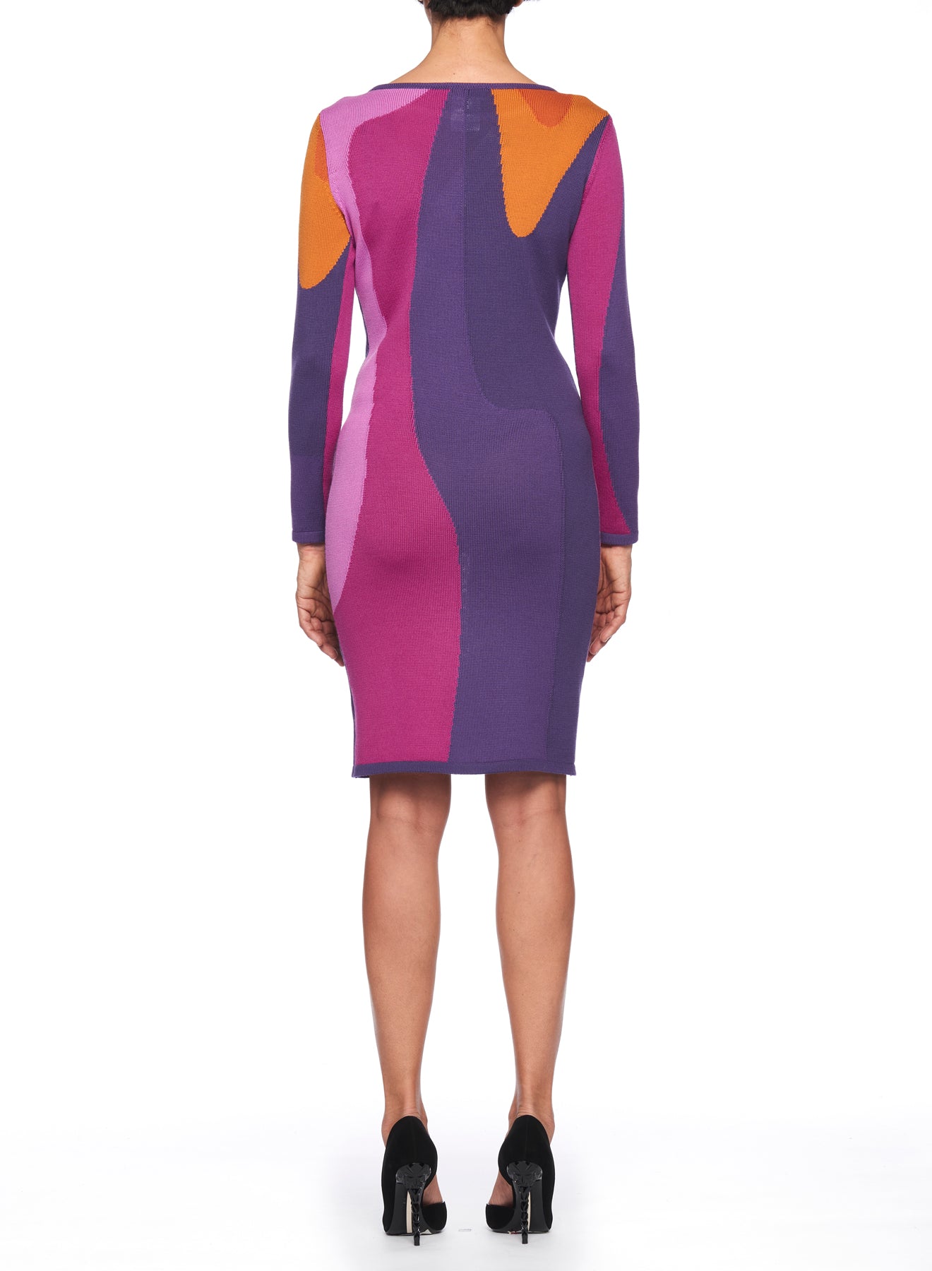 A person is wearing the Fuzzi Woman Abstract Art Dress by Fuzzi, featuring knee-length, multicolored patterns in purple, pink, orange, and red that resemble vibrant color-blocked art. They stand against a white background in black high-heeled shoes. The dress has long sleeves.