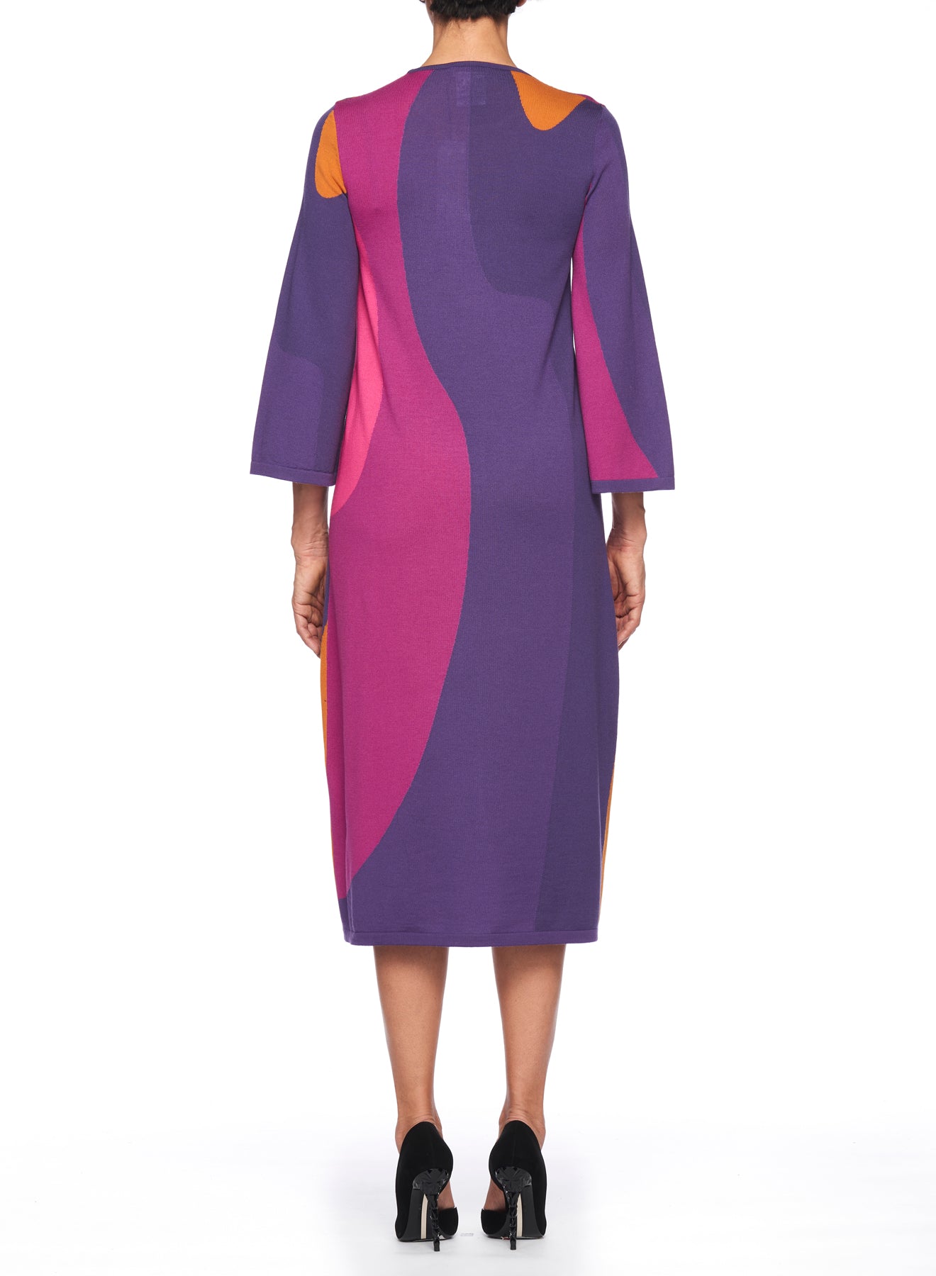 A person wearing the Fuzzi Woman Retro Vibes Dress stands facing away. This mid-length dress, created by Fuzzi, features a vibrant abstract pattern in shades of purple, pink, and orange, evoking a vintage charm. Its three-quarter sleeves pair perfectly with their black heeled sandals.