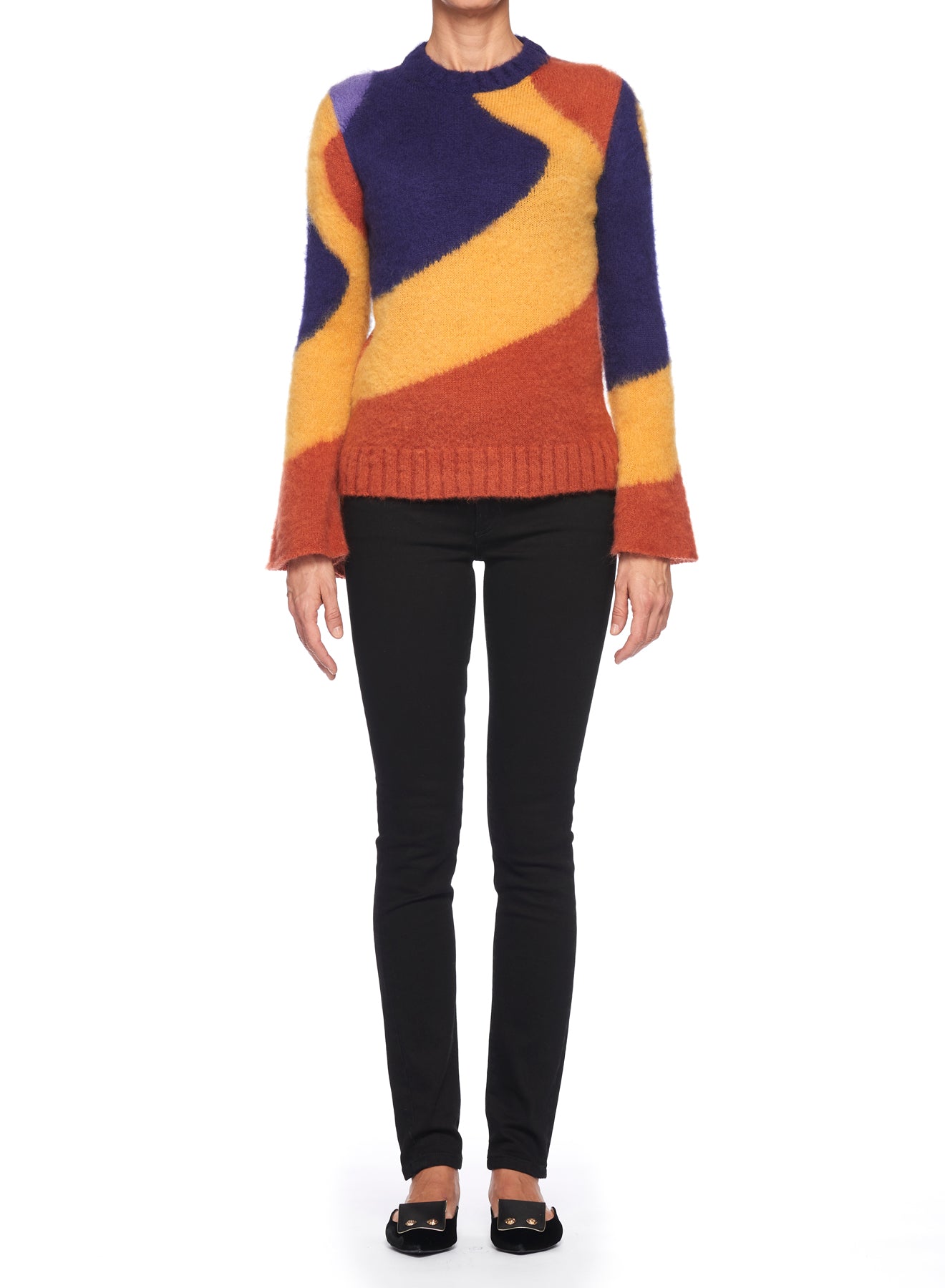 The individual is dressed in a vibrant Fuzzi Woman Colorful Sweater by Fuzzi, featuring shades of yellow, orange, and purple. They are paired with black pants and black shoes. The plain white background highlights their retro-inspired ensemble beautifully.