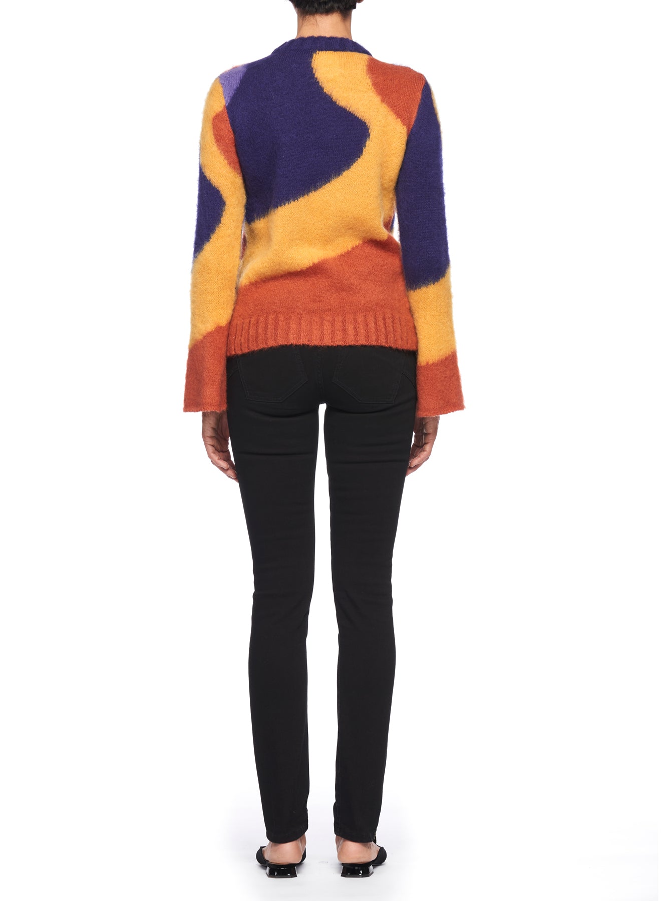 A person dressed in the Fuzzi Woman Colorful Sweater in vibrant hues of orange, purple, and yellow, complemented by black skinny jeans, stands against a plain white background. The image showcases the back view, highlighting a retro-inspired look from the Fuzzi brand.