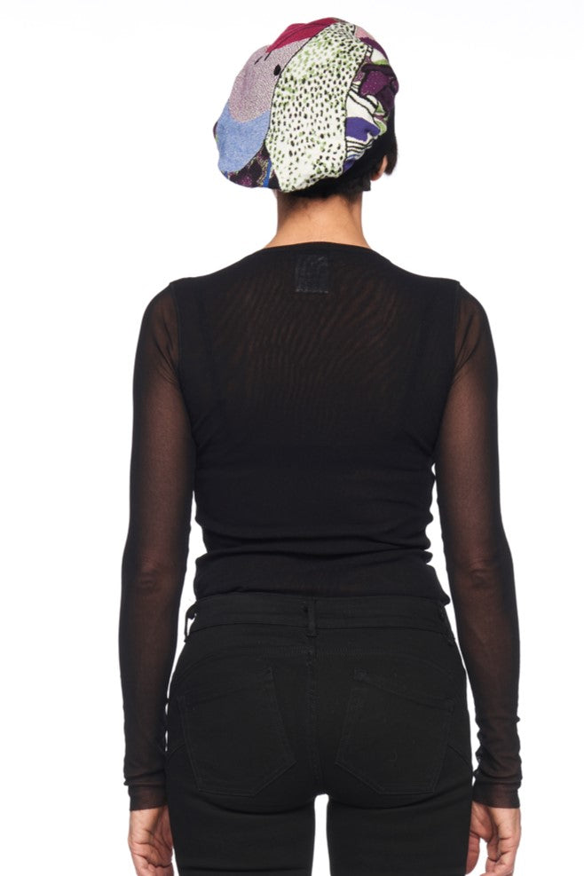 A person is seen from behind, wearing a sheer black long-sleeve top and black jeans, perfectly complemented by the Fuzzi Woman Beanie—a charming vintage piece featuring colorful shades of purple, green, and white—set against the plain white backdrop.