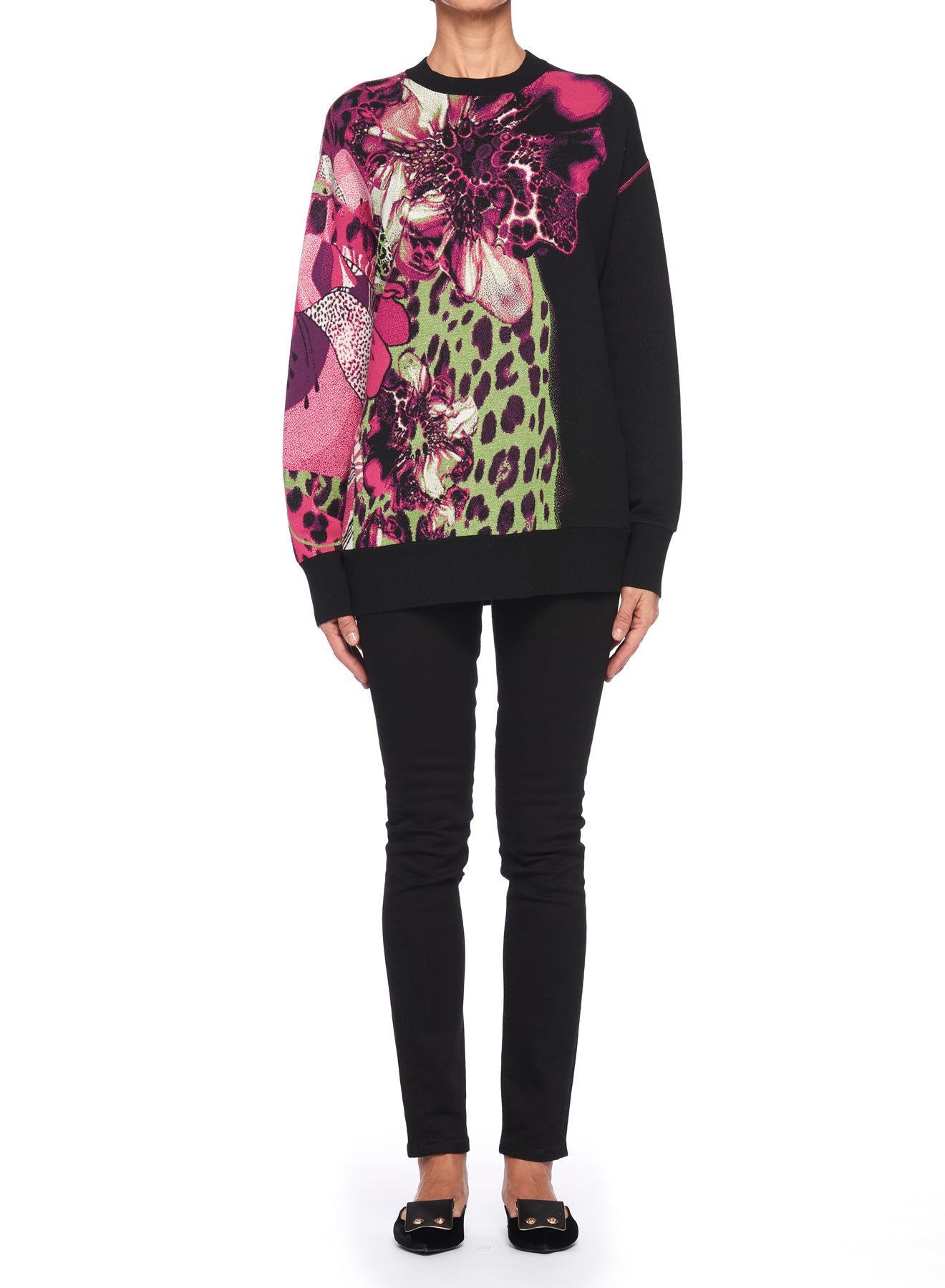 A person stands wearing the Fuzzi Woman Bold and Edgy Graphic Sweater by Fuzzi, characterized by its oversized fit and vibrant floral and leopard print designs in pink, green, and black. They complete the outfit with black pants and black shoes against a plain white background.