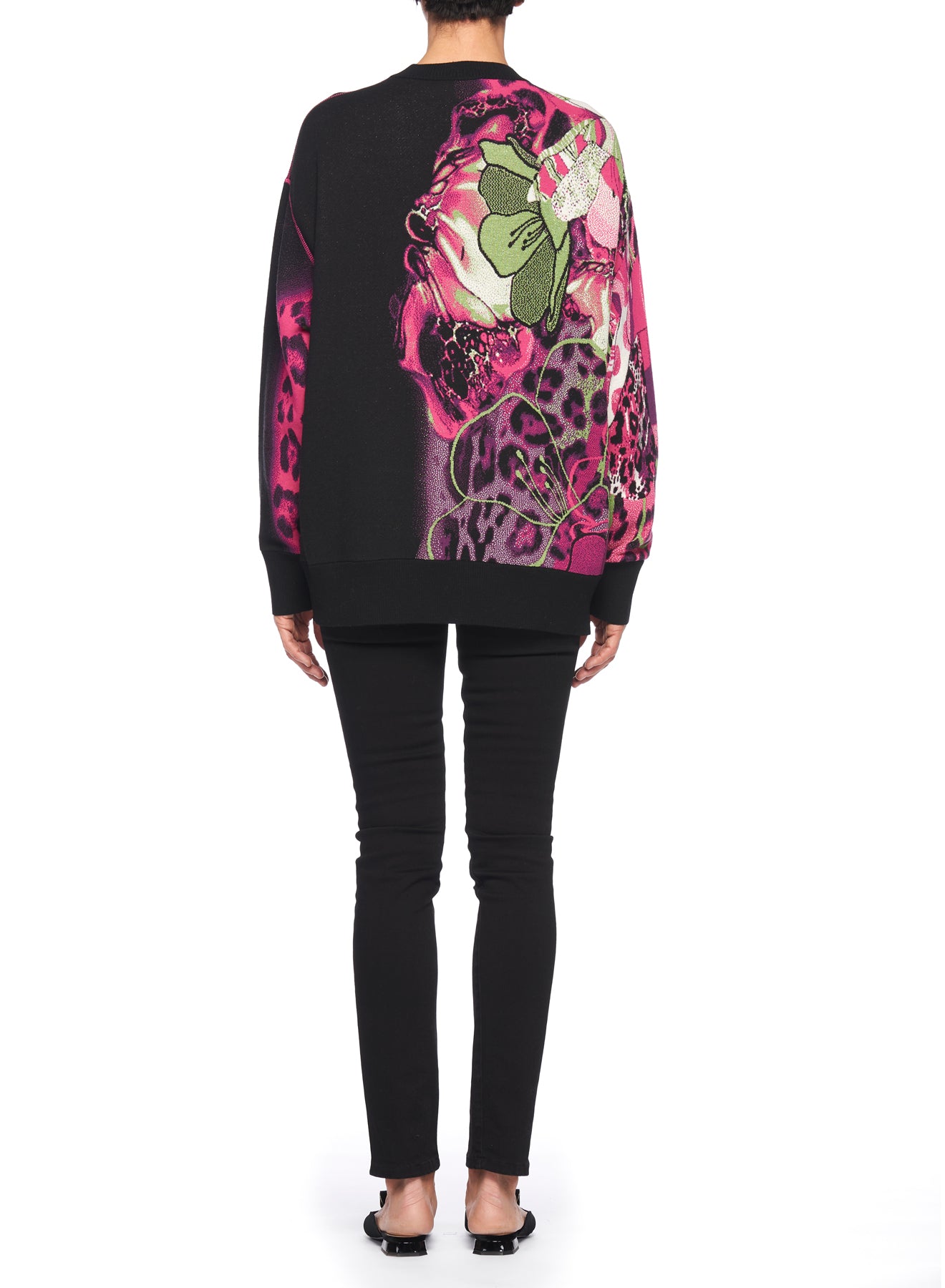 A person wearing the Fuzzi Woman Bold and Edgy Graphic Sweater, boasting vibrant floral patterns in pink and green tones, paired with black pants. They are standing against a plain white background, facing away from the camera.