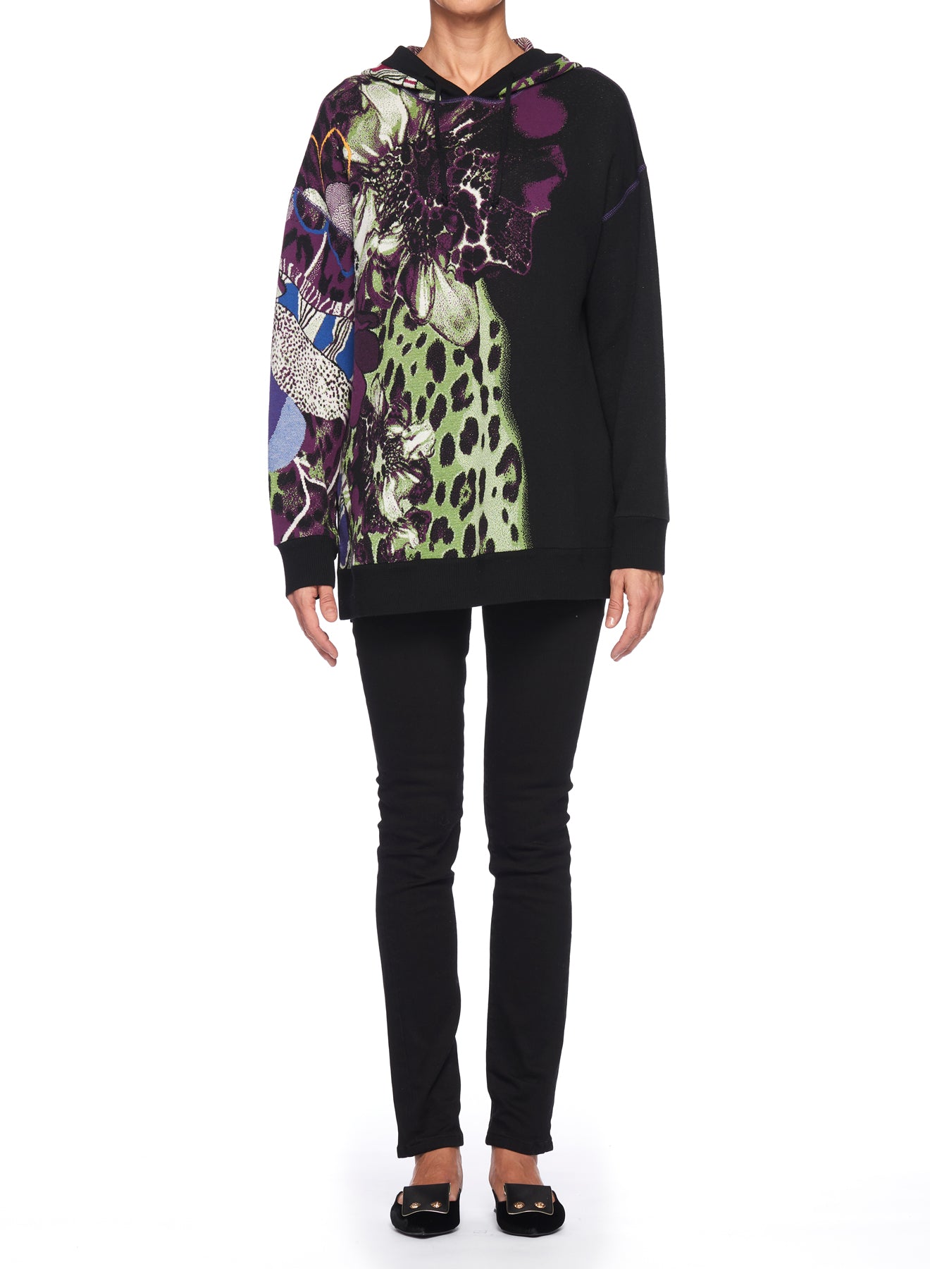 A person wearing a vintage piece—the Fuzzi Woman Graphic Hoodie featuring vibrant, psychedelic floral and animal print patterns in purple, green, and black—stands against a white background. The oversized fit pairs seamlessly with black pants and shoes, showcasing a unique blend of textures and colors.