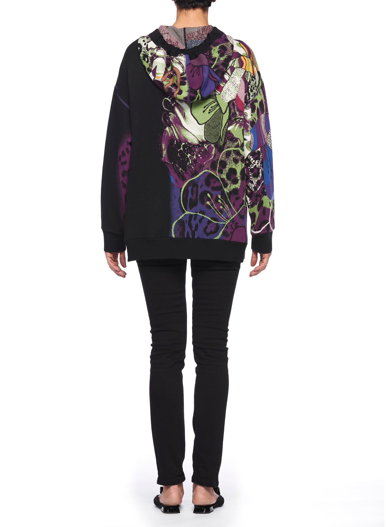 A person stands with their back to the camera, wearing the Fuzzi Woman Graphic Hoodie by Fuzzi. This black hoodie features a vibrant abstract design of flowers and leopard spots. The oversized fit effortlessly complements black pants and flats, all set against a plain white background.