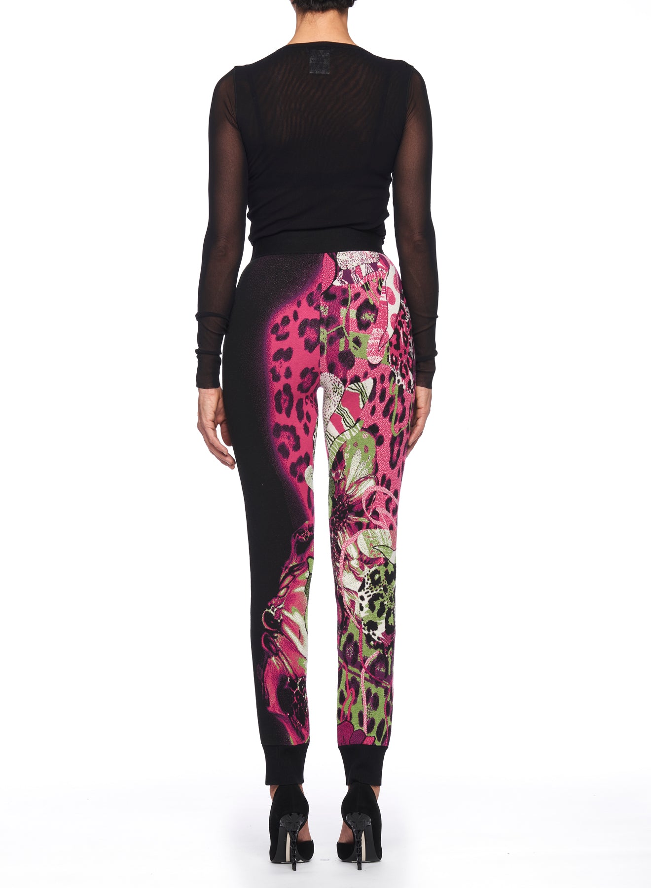 A person is wearing a black sheer long-sleeve top paired with Fuzzi Woman Wild Side Joggers, featuring a mix of pink, black, and green prints. Complemented by their vintage piece high-heeled shoes, the photo captures their ensemble from behind against a white background.