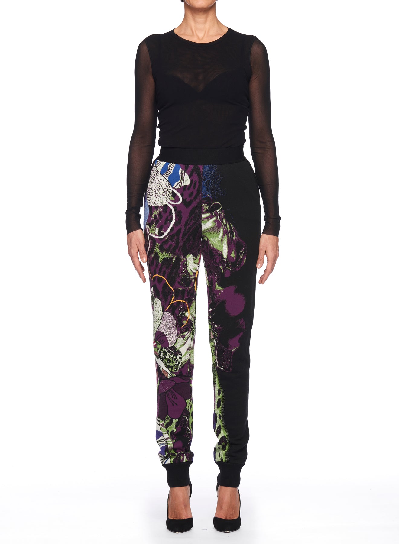 A person wearing a sheer black long-sleeve top and the Fuzzi Woman Wild Side Joggers by Fuzzi stands against a plain white background. The joggers showcase abstract designs in purple, green, and other shades, making them a striking vintage piece.